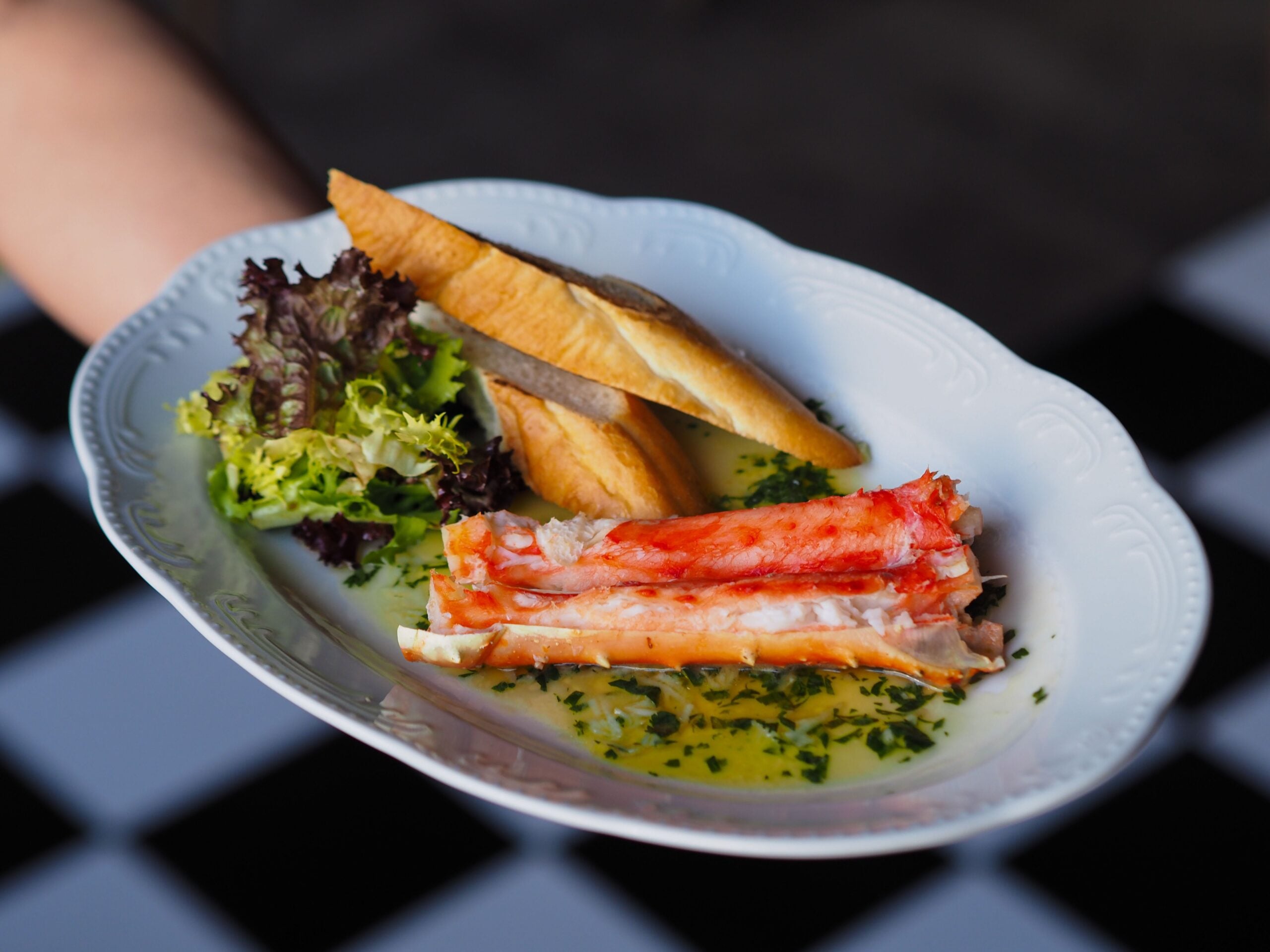 A white plate with crab legs in herb butter, two slices of toasted bread, and a small side of mixed greens, held by a hand over a black and white checkered surface.
