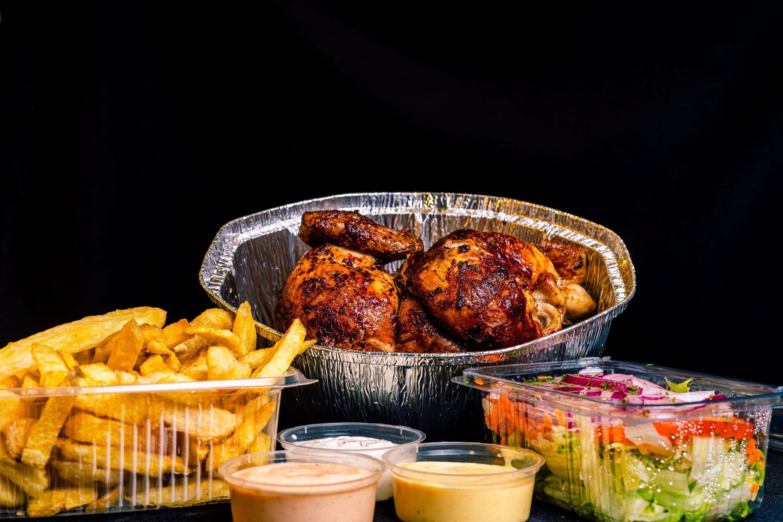 A meal of roasted chicken in a foil container, served with French fries, a fresh salad, and dipping sauces, all set against a black background.
