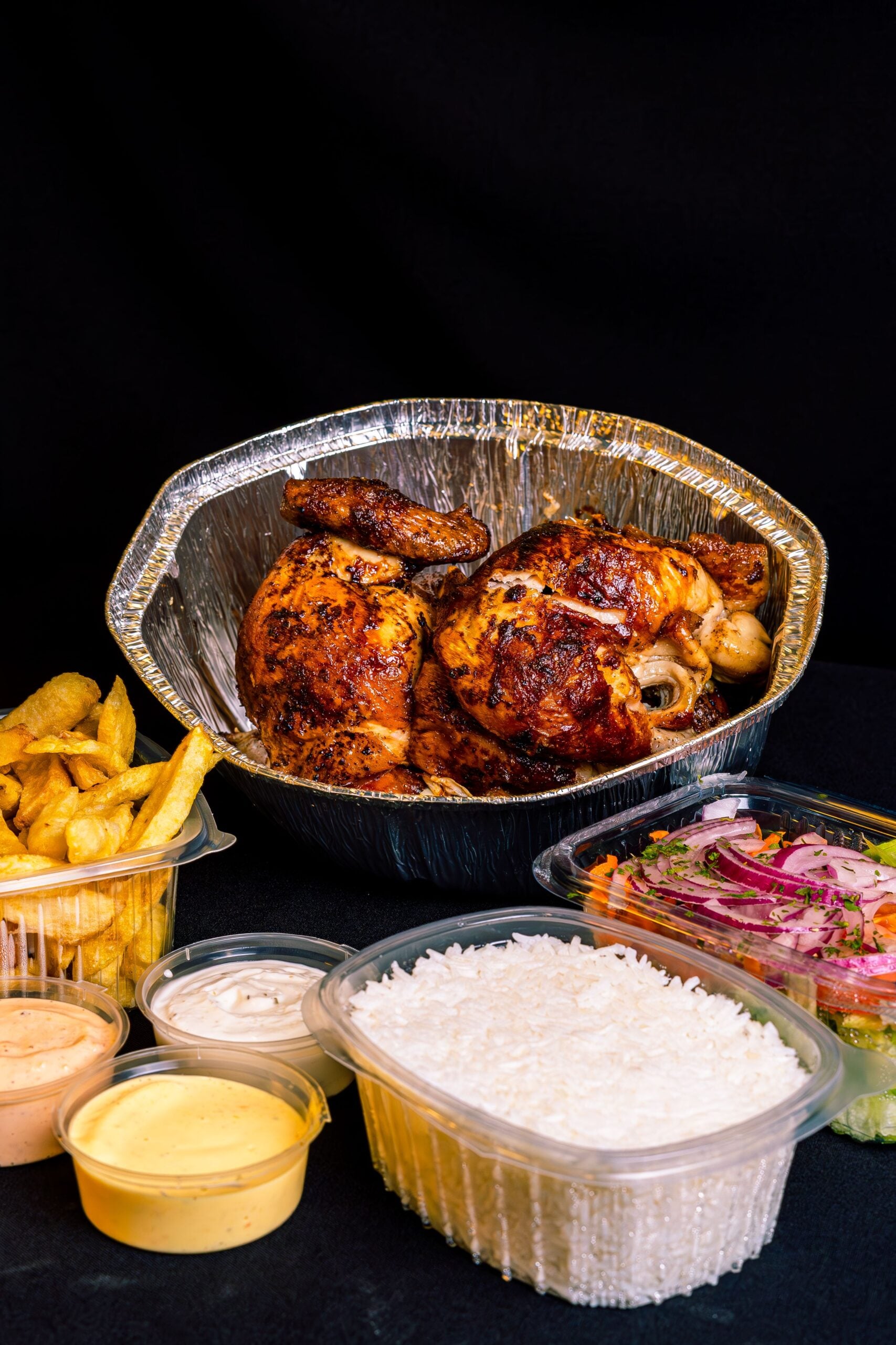 A meal with roasted chicken in a foil container, white rice, golden fries, fresh salad with onions, and three dipping sauces, all set against a black background.