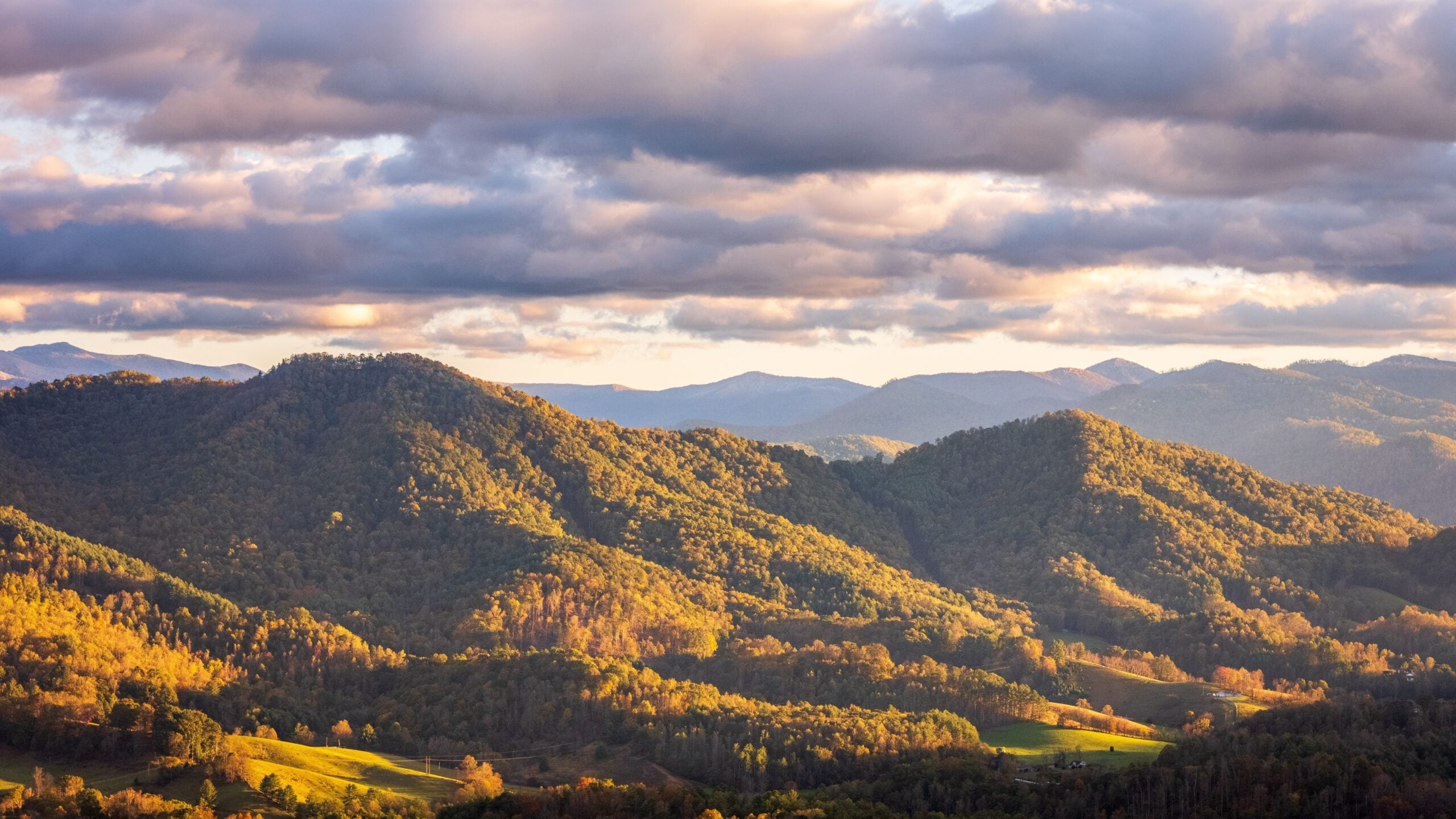 A scenic view of rolling, tree-covered mountains under a partly cloudy sky, with sunlight casting warm, golden highlights on the landscape and patches of green fields in the valleys below.
