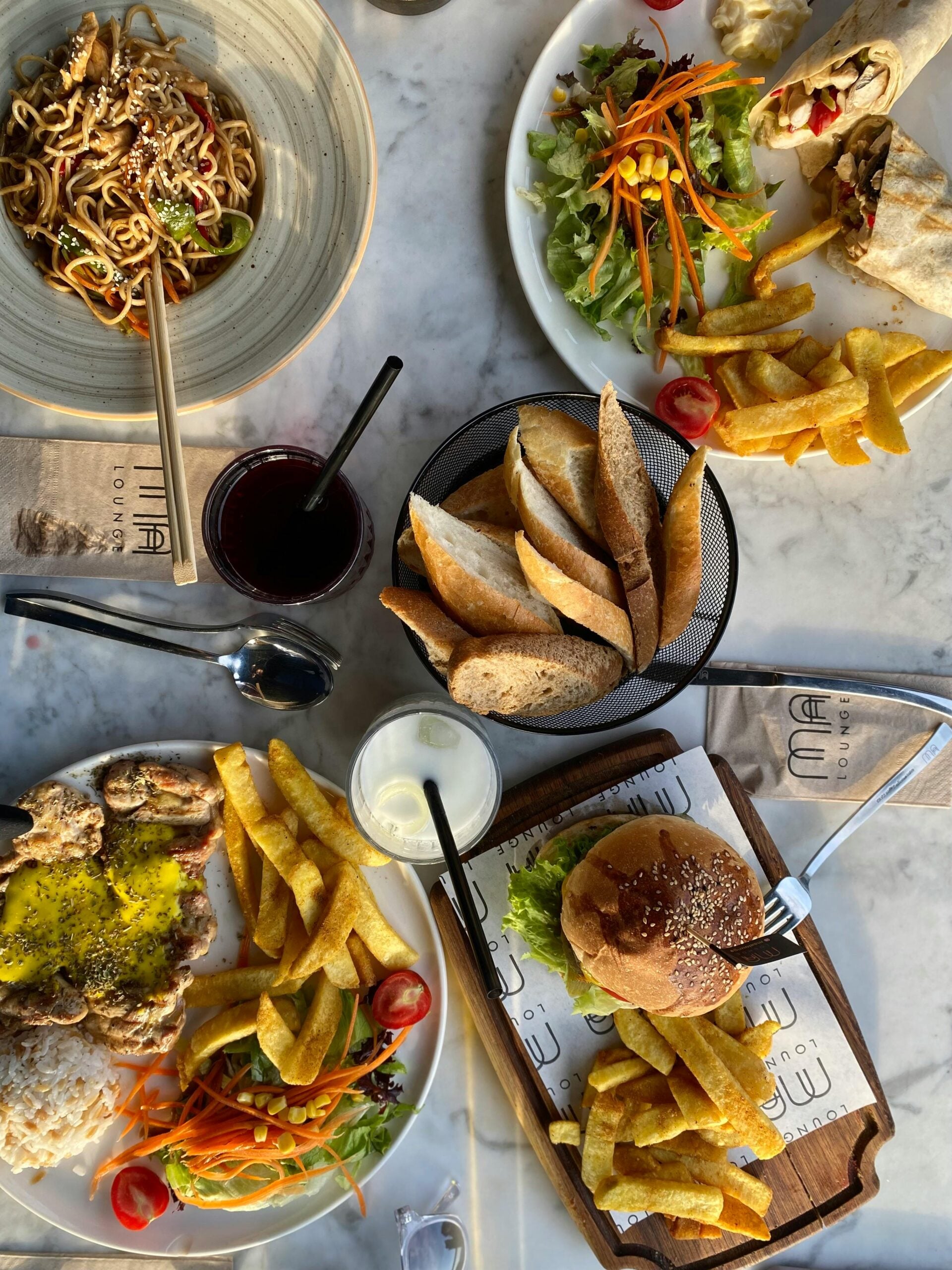 A table set with a variety of meals including a noodle dish, a wrap with fries and salad, a bread basket, grilled chicken with fries and rice, a burger with fries, drinks, and garnished vegetables on a marble surface.