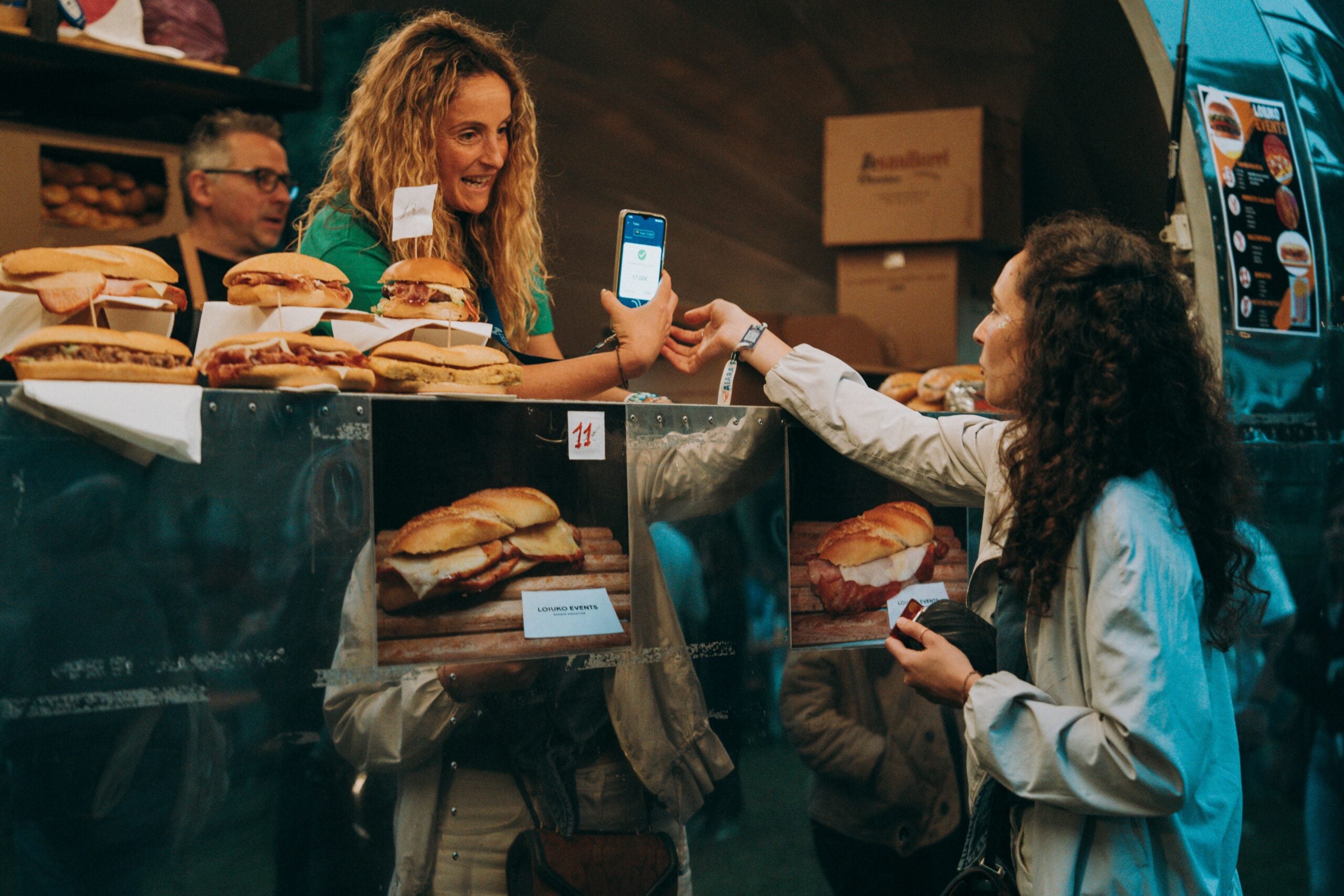 A woman behind a food stall counter smiles while holding up a phone for payment; another woman reaches out to scan it, surrounded by sandwiches and menu photos. Other people are visible in the background.