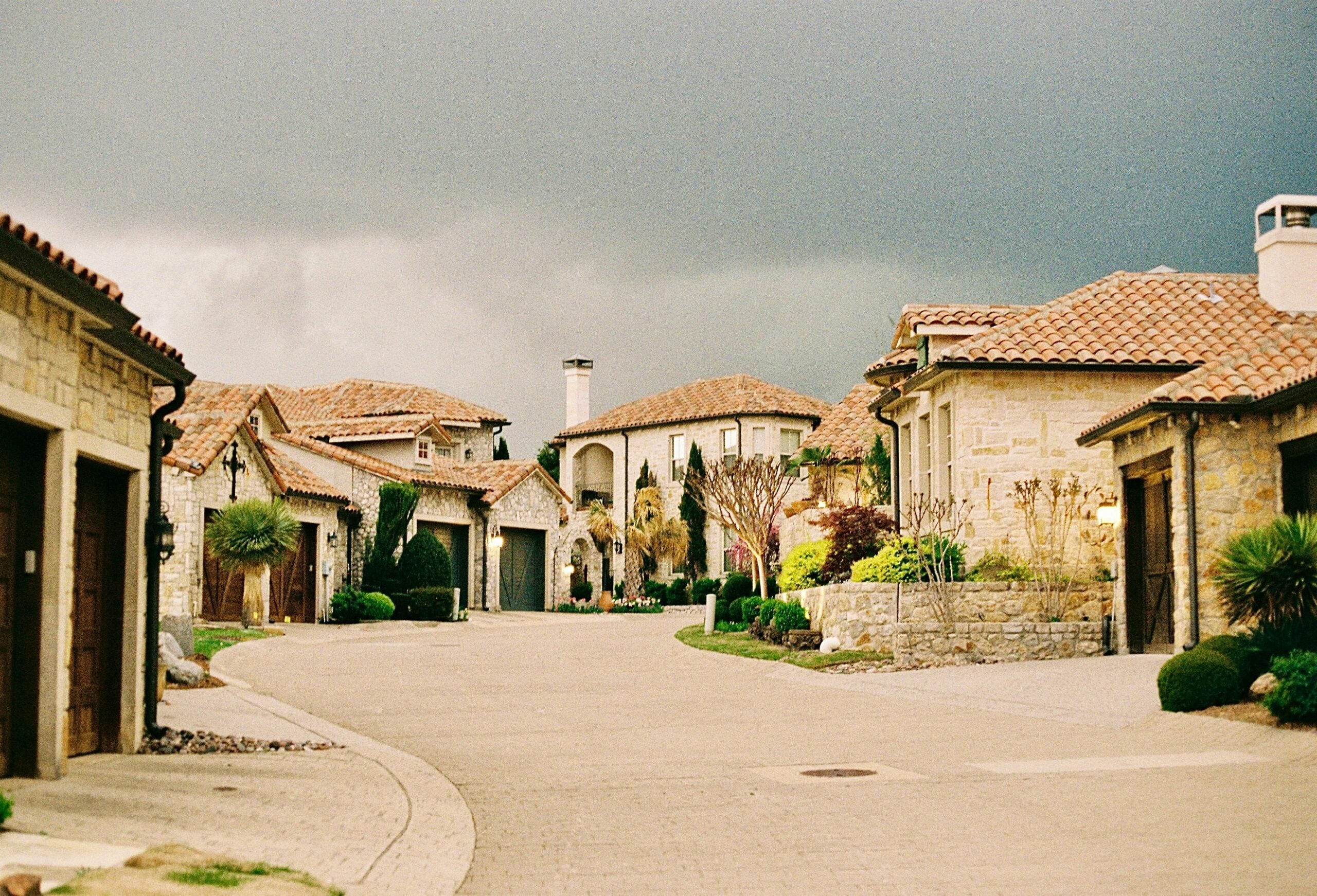 A quiet residential street with stone houses and red-tiled roofs under a cloudy, dark gray sky; well-kept gardens and greenery line the curved road.
