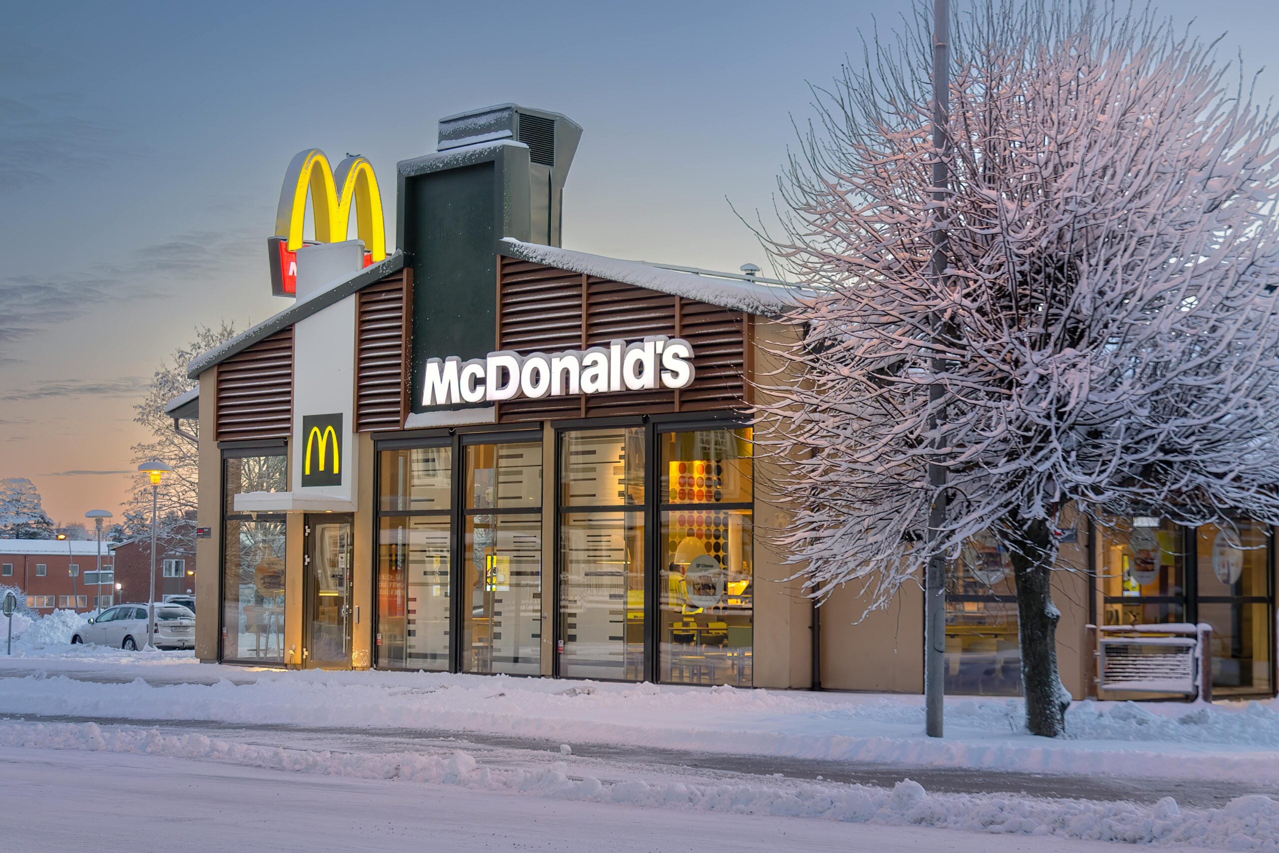 A McDonald's restaurant with large illuminated signs is surrounded by snow-covered trees and ground, captured during winter at dusk. The interior lights create a warm contrast to the cold outdoor scene.