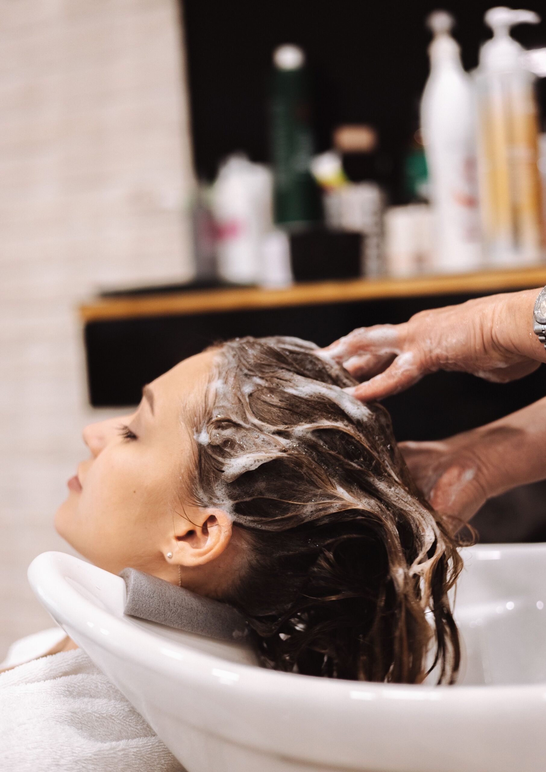A woman reclines at a salon sink with her eyes closed while a hairdresser washes her hair, creating a lather of shampoo. Bottles of hair products are blurred in the background.