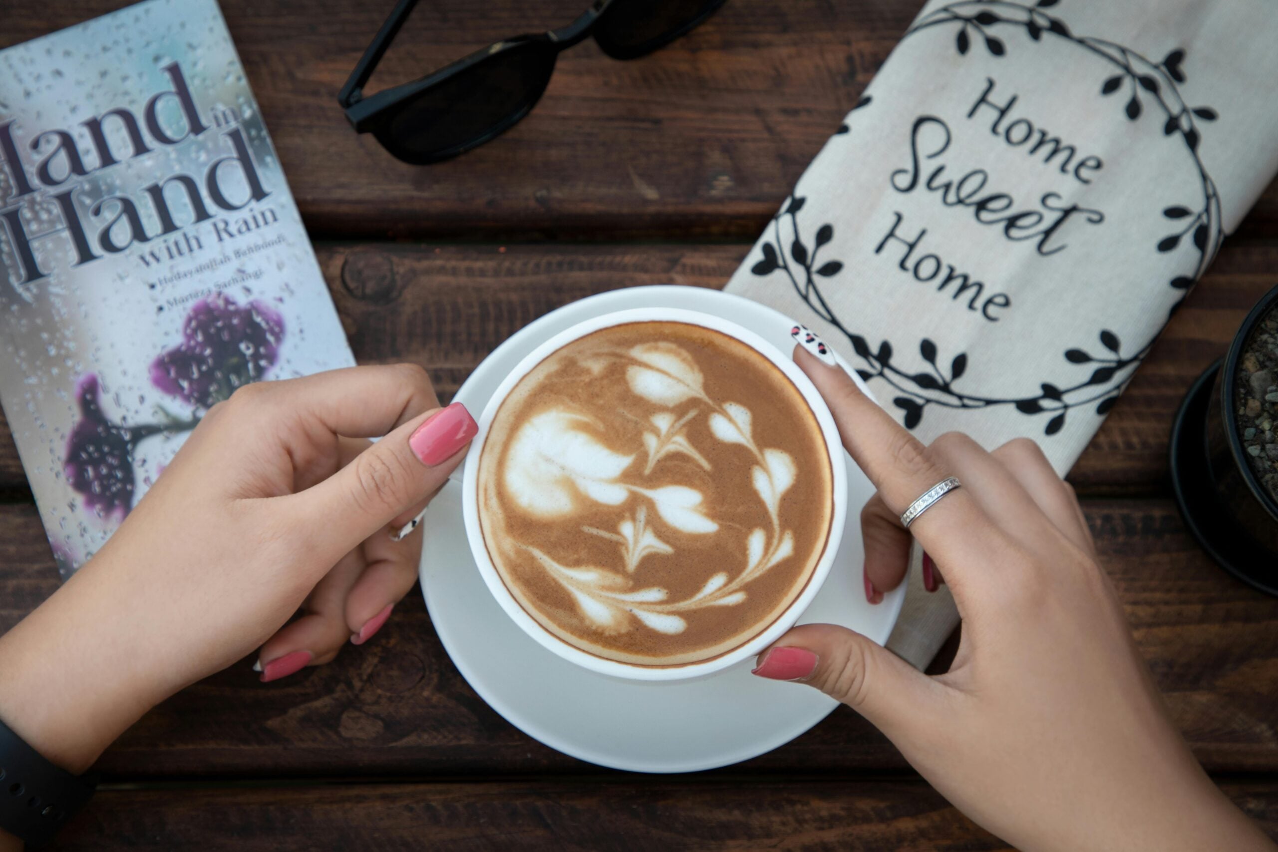 A person with pink nails holds a cup of latte with intricate leaf latte art, next to a "Home Sweet Home" cloth, sunglasses, a book, and a plant on a wooden table.