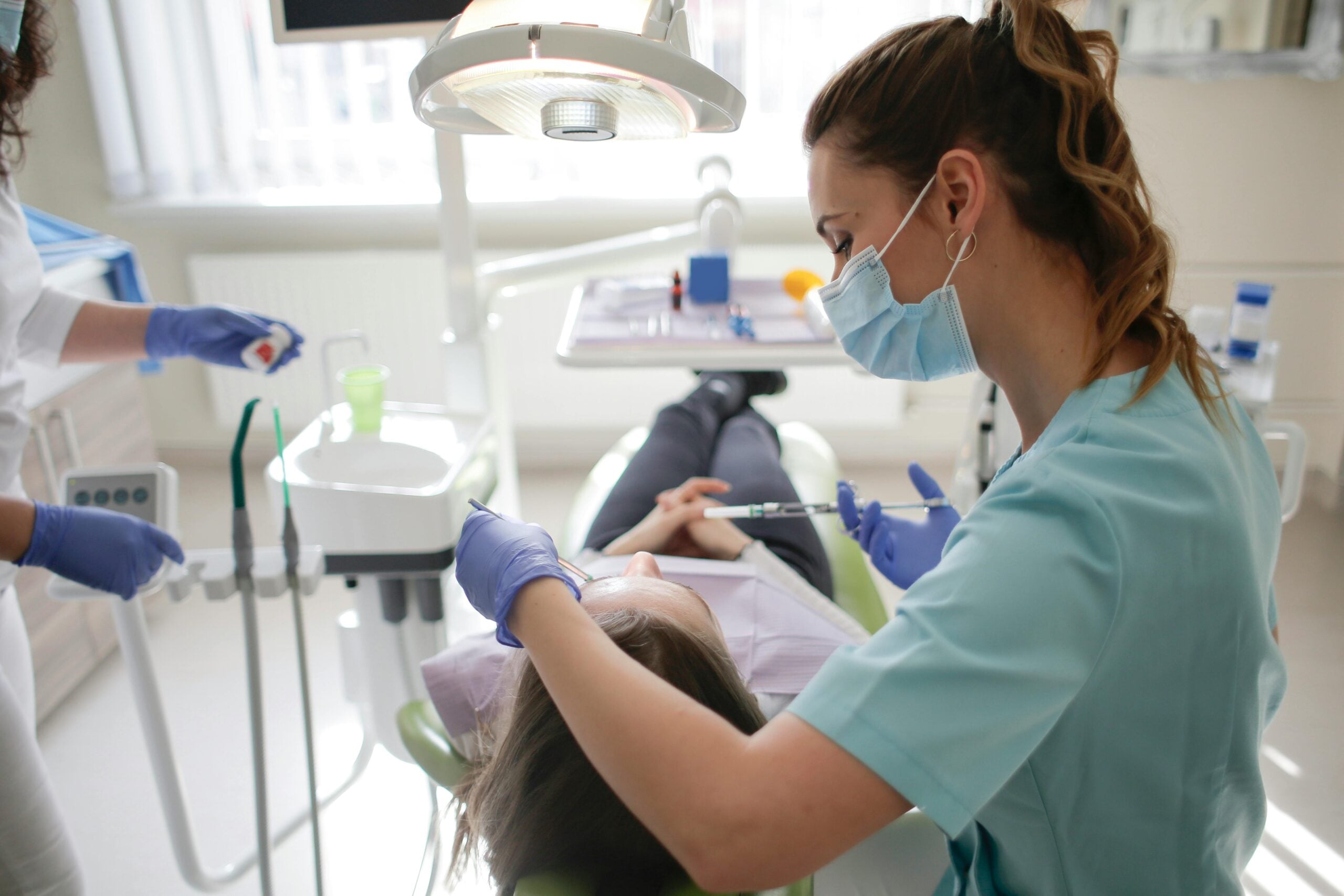 A dentist wearing a mask and gloves examines a patient lying in a dental chair, while an assistant stands nearby holding a tool in a bright dental clinic.