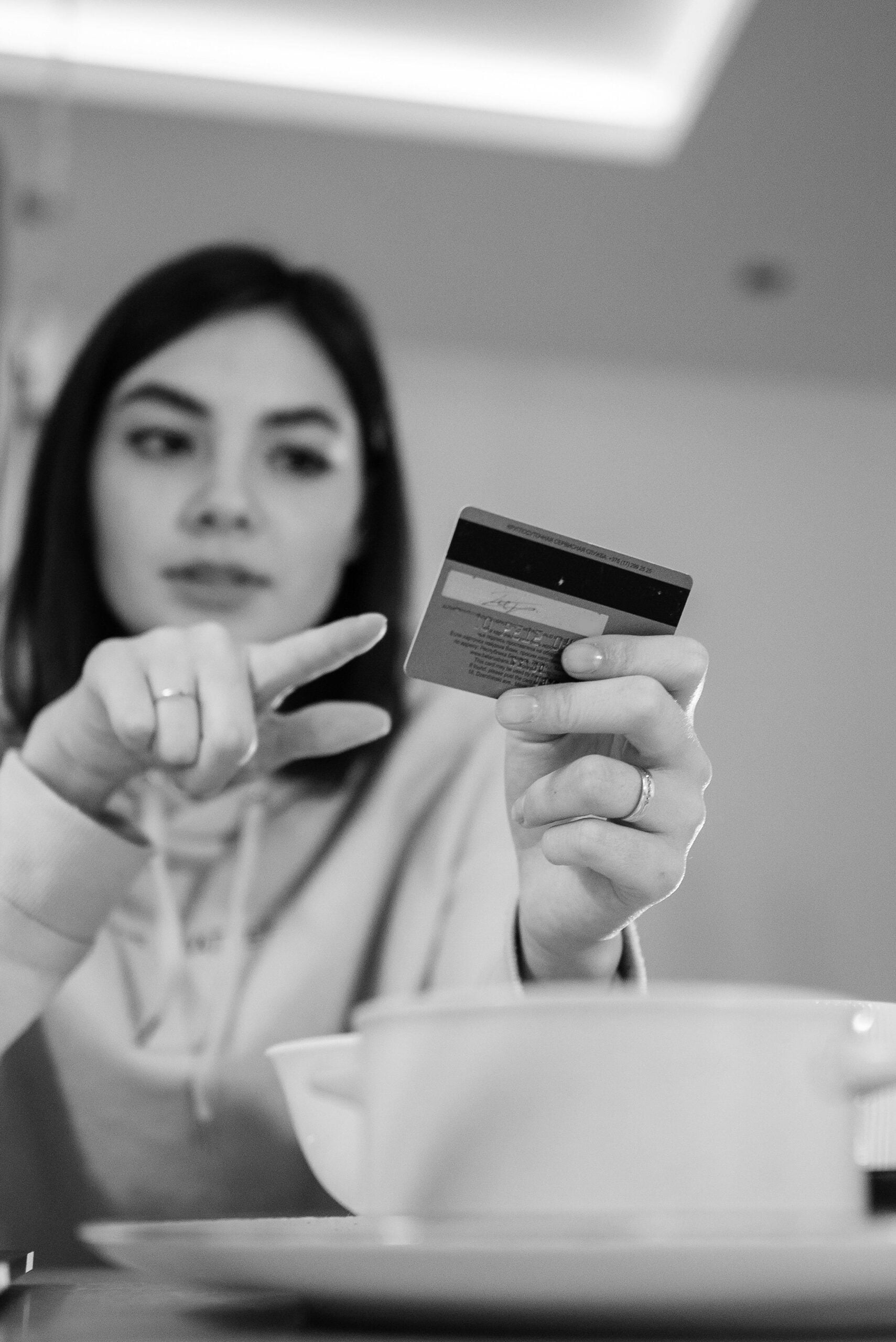 A woman holding a credit card in one hand and pointing at it with the other, sitting at a table with a bowl in front of her. The image is in black and white.