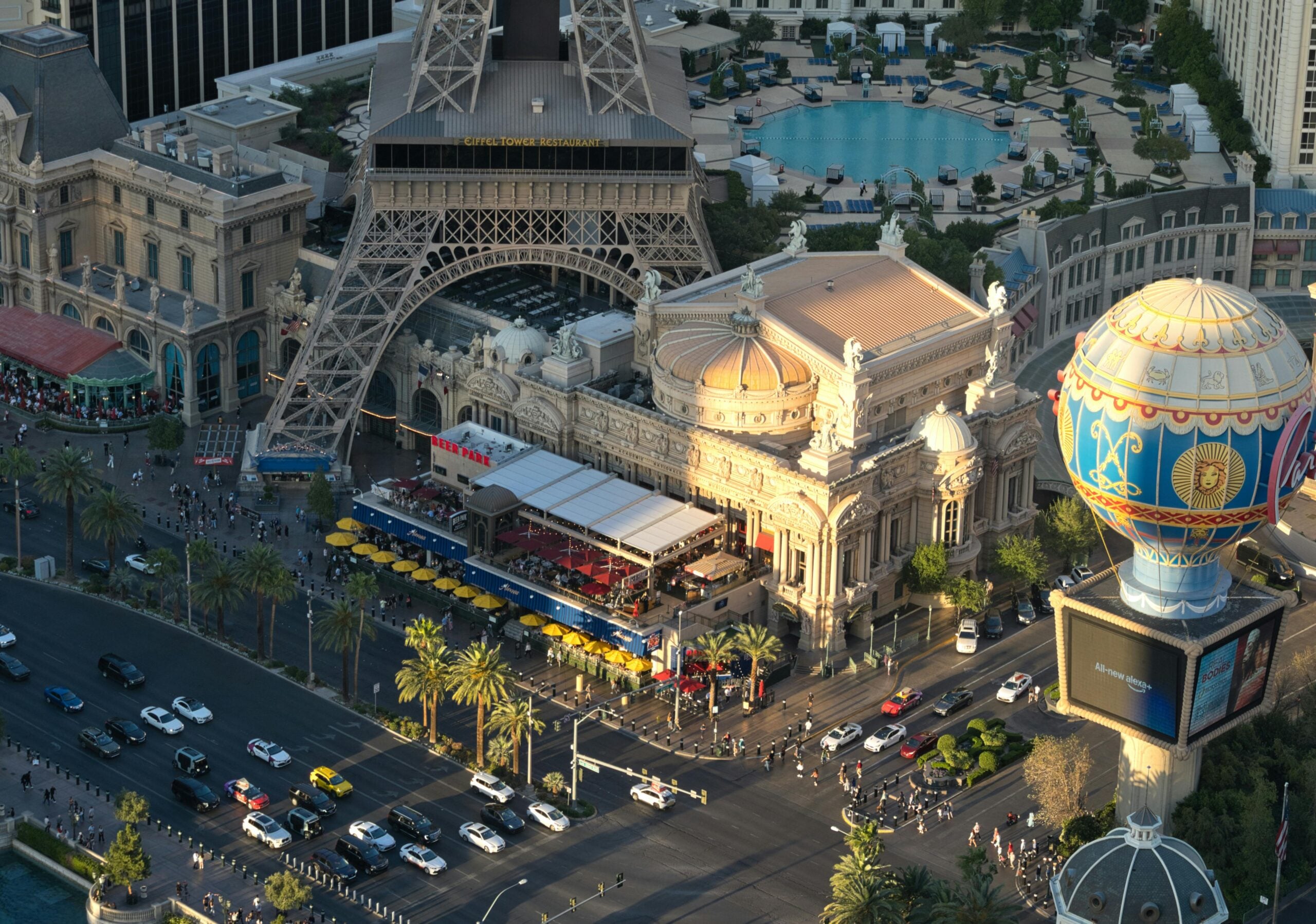 Aerial view of the Paris Las Vegas Hotel featuring a replica Eiffel Tower, ornate buildings, a pool, a hot air balloon sign, and busy streets filled with pedestrians and vehicles.