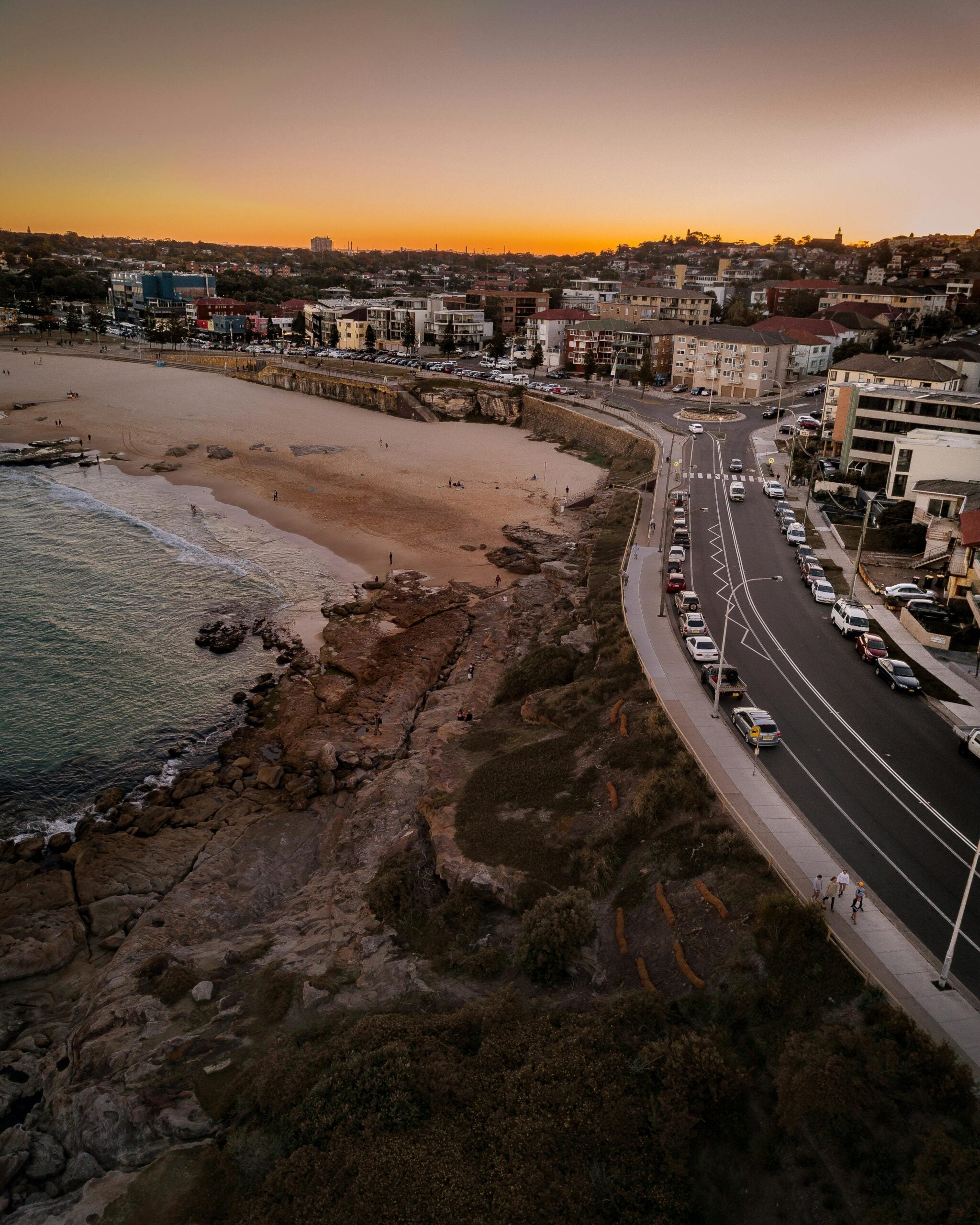Aerial view of a beach at sunset with golden light over sand, rocky shoreline, parked cars along a coastal road, and buildings in the background.