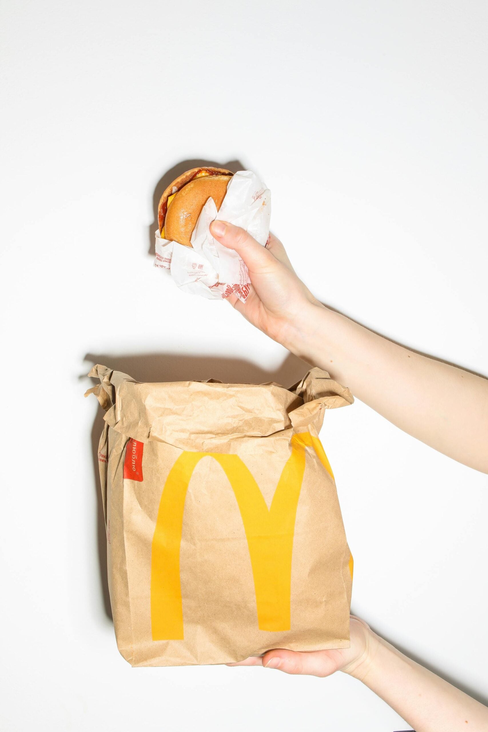 A person holds a wrapped McDonald's burger above a brown McDonald's paper bag with the yellow logo, against a plain white background.
