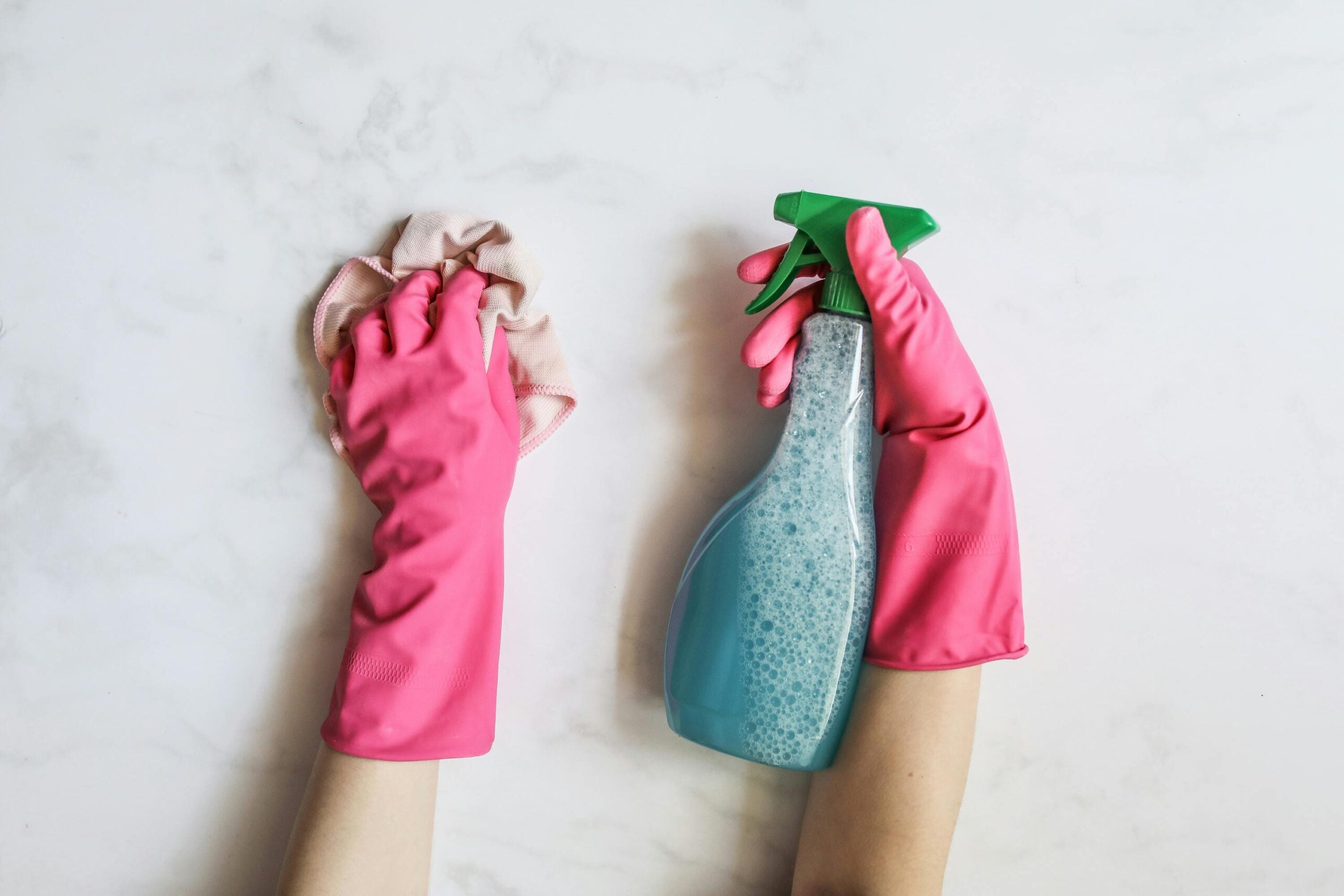 Hands wearing pink rubber gloves hold a spray bottle with blue liquid in one hand and a cleaning cloth in the other, cleaning a white surface.