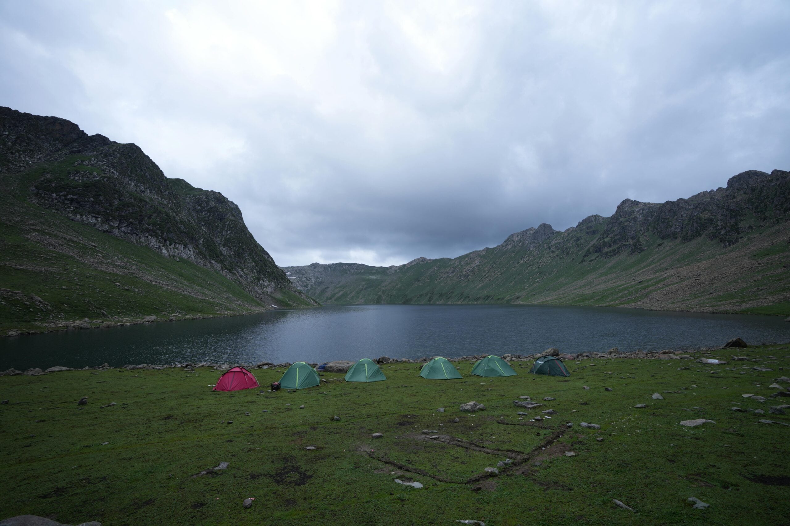 Several colorful tents are set up on grassy ground beside a tranquil lake, surrounded by green mountains under a cloudy sky.