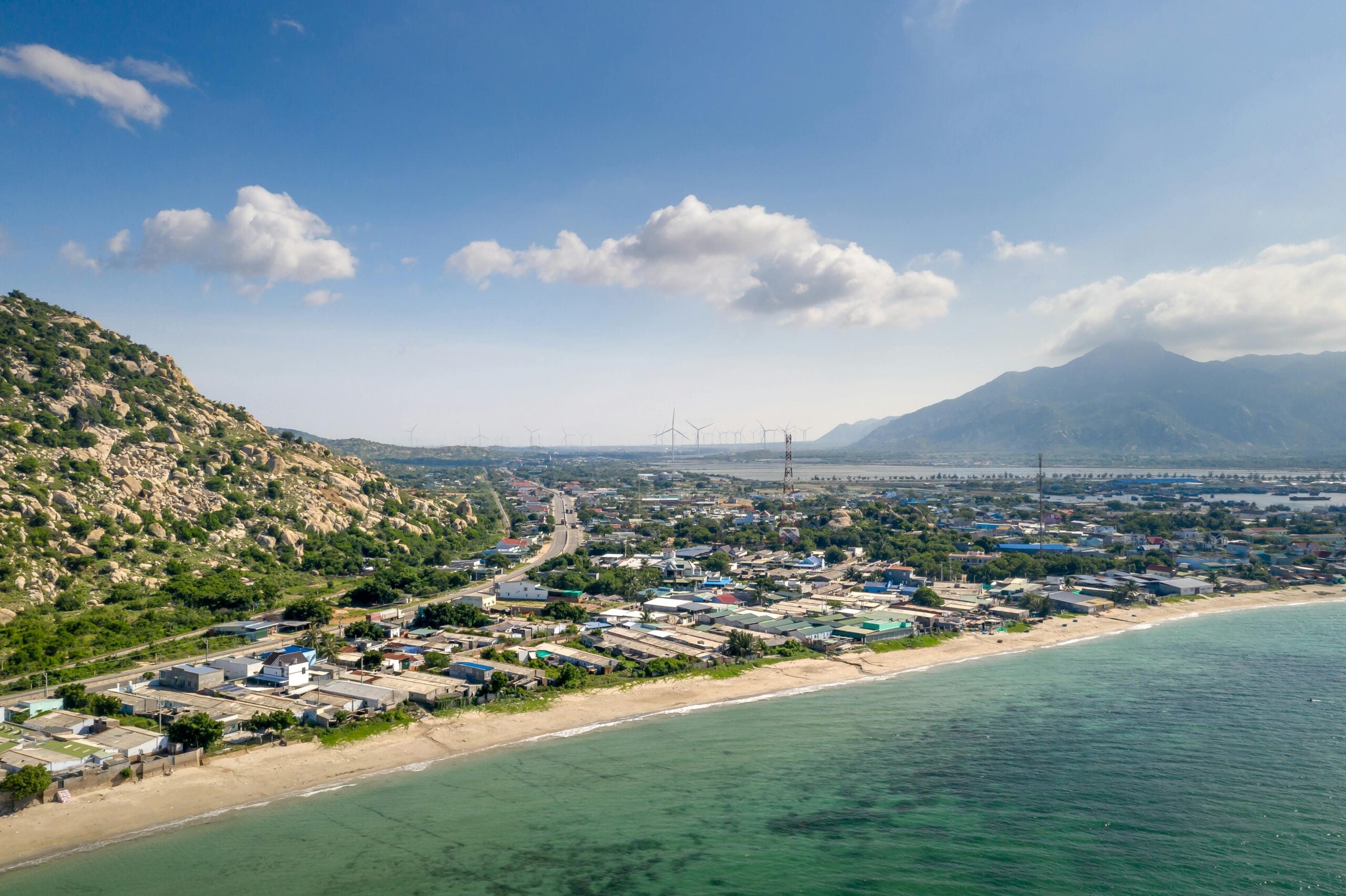 A coastal town with sandy beaches, clear turquoise water, and green hills. Houses and buildings line the shore, and wind turbines are visible in the distance beneath a partly cloudy sky.