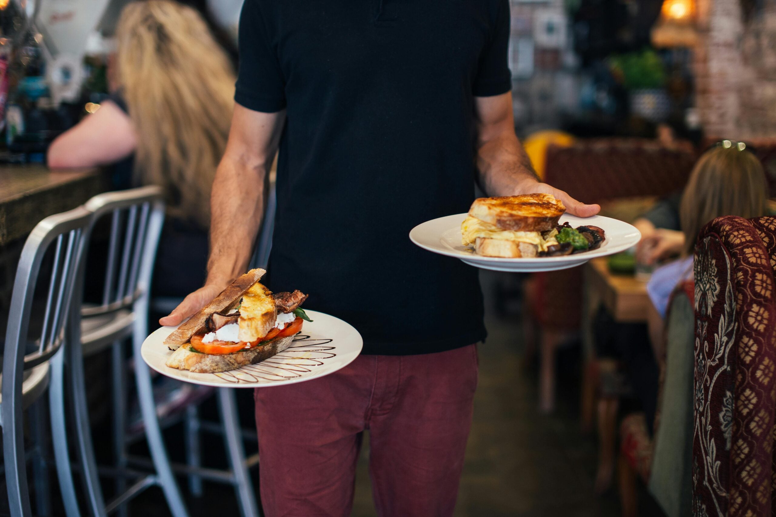 A server in a black shirt and red pants holds two plates of food in a busy restaurant. One plate has a sandwich with tomato, lettuce, and meat; the other plate has a sandwich and salad.