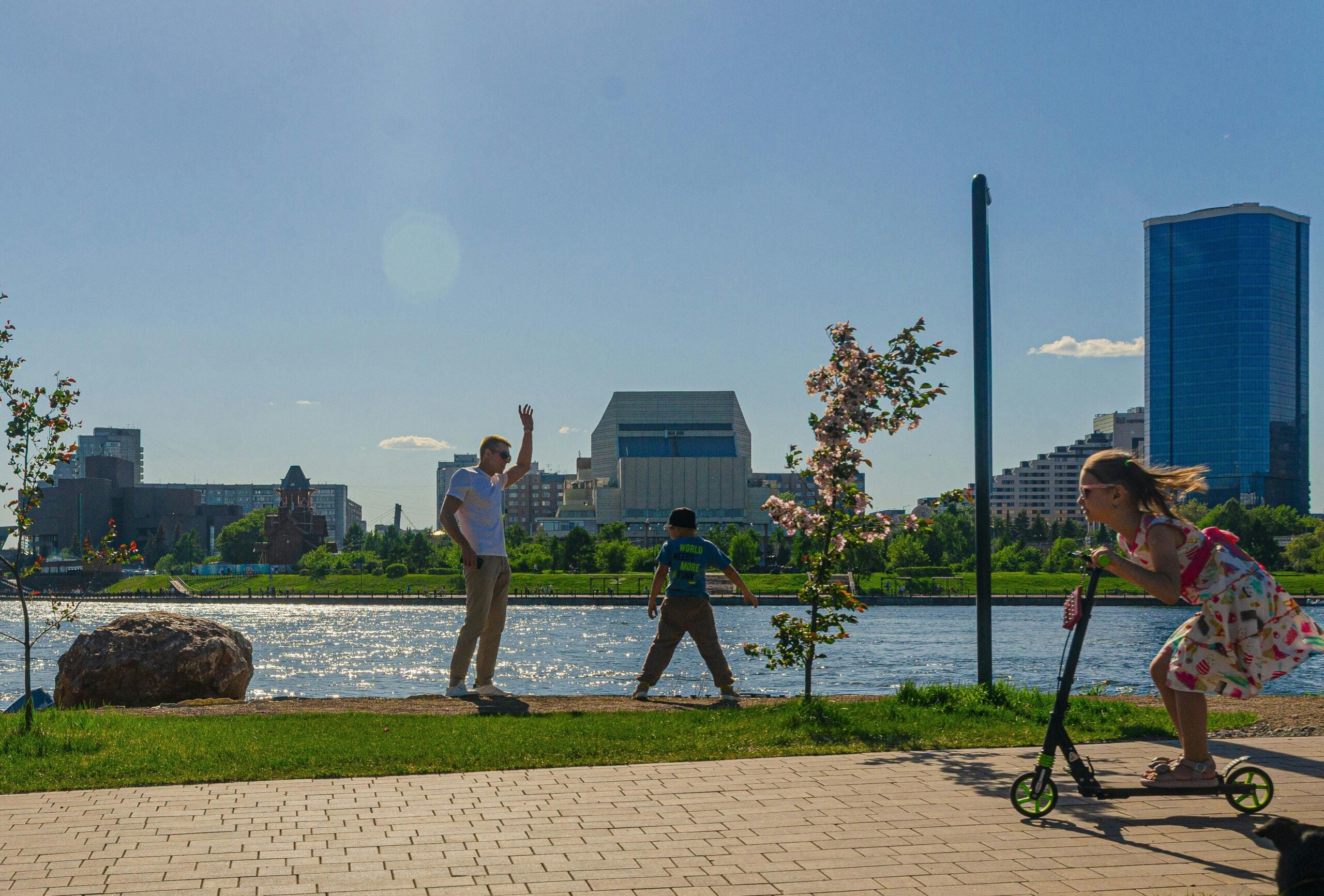 A girl rides a scooter on a riverside path while two people, one adult and one child, play near the water. City buildings and trees are visible in the background under a clear blue sky, reflecting the benefits of Cost of Living Relief in daily life.