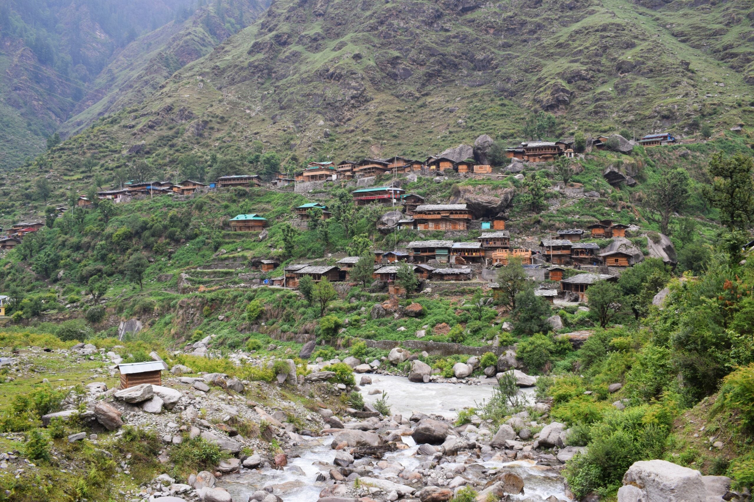 A cluster of wooden houses is spread across a lush green hillside above a rocky river, surrounded by mountains and dense vegetation.