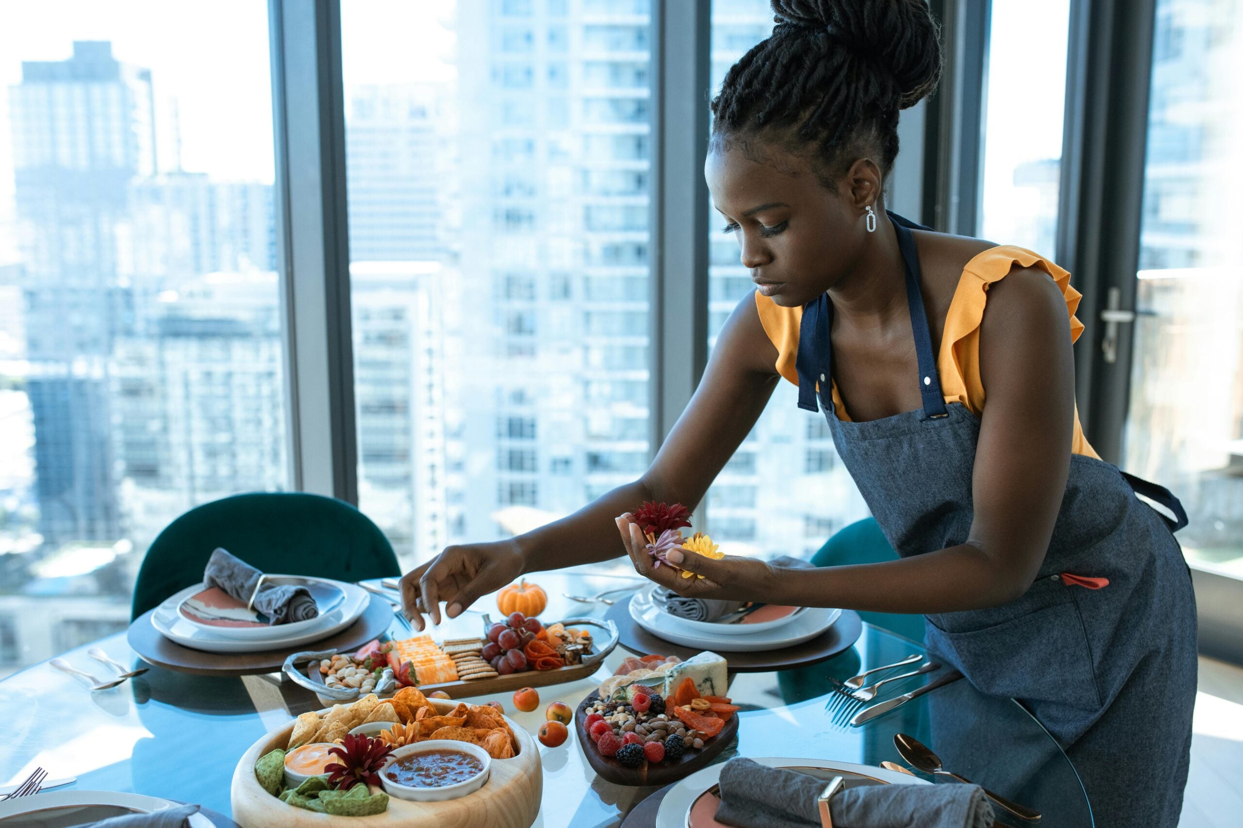 A woman in an apron arranges fruit and cheese on a platter at a dining table set with plates, napkins, and cutlery, in a bright room with large windows overlooking a cityscape.