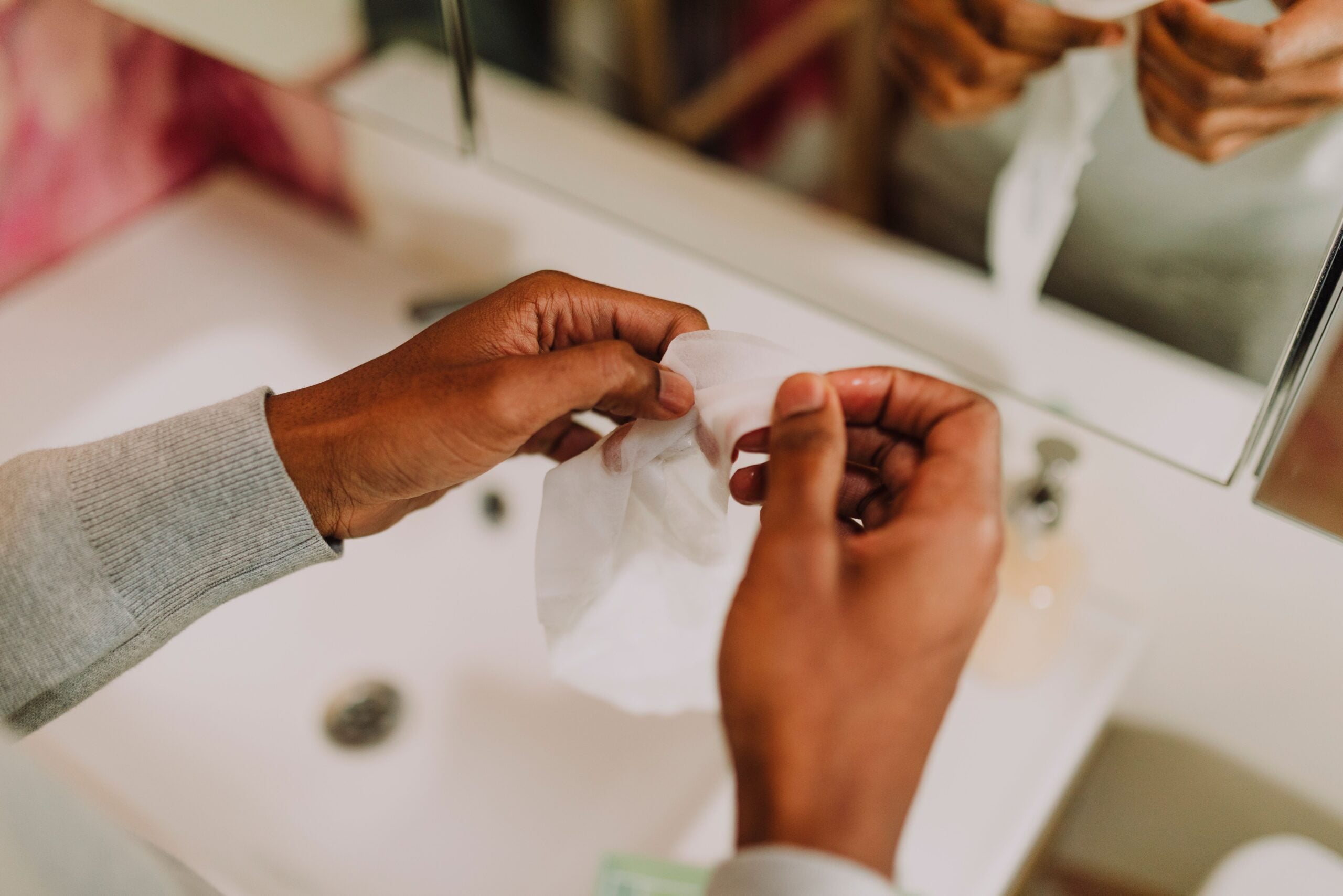 A person holding a white wet wipe over a bathroom sink, with a mirror and various items blurred in the background.