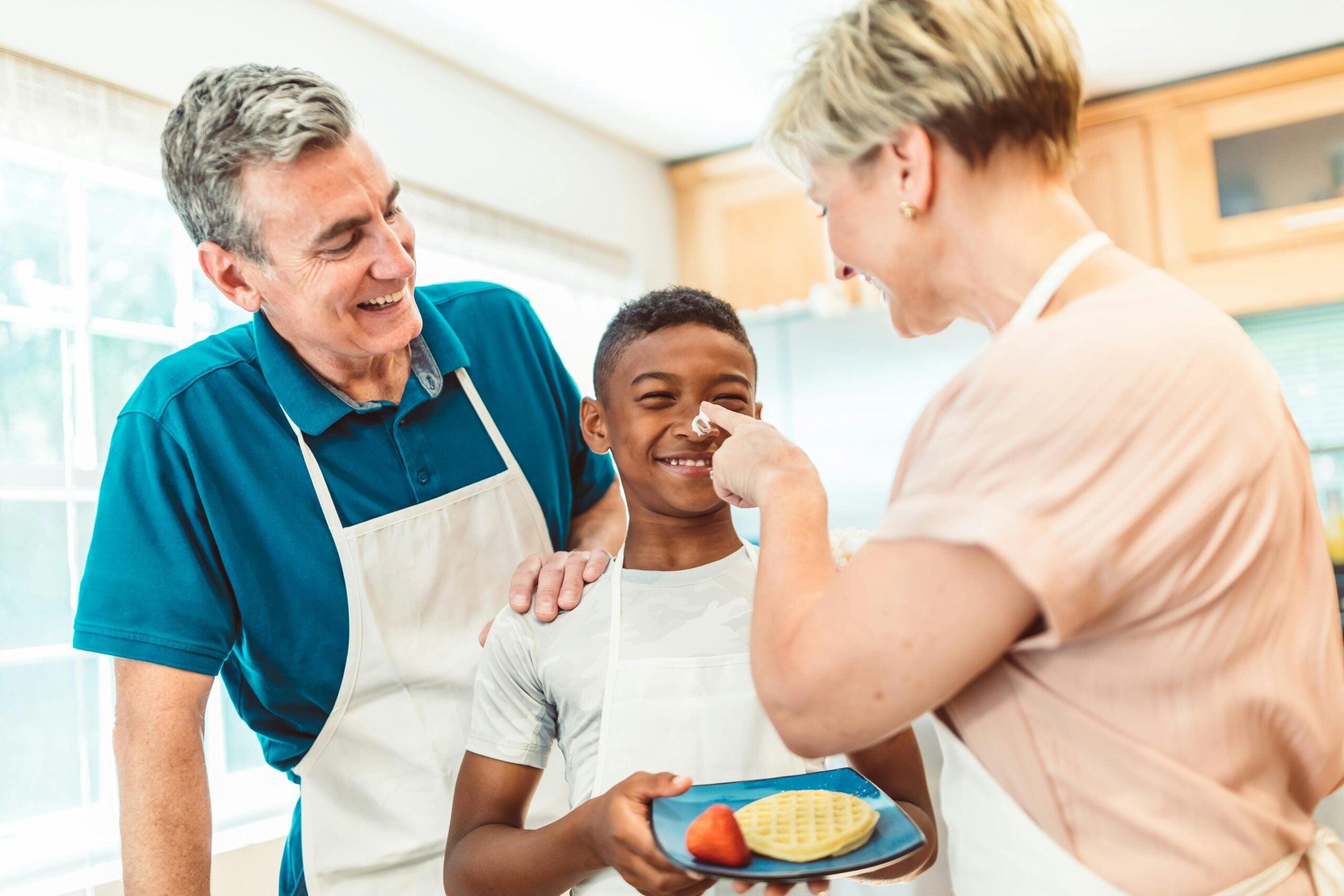 A smiling child holds a plate with a waffle and strawberry while an adult woman playfully puts whipped cream on his nose. An older man, also smiling, stands beside them in a bright kitchen.