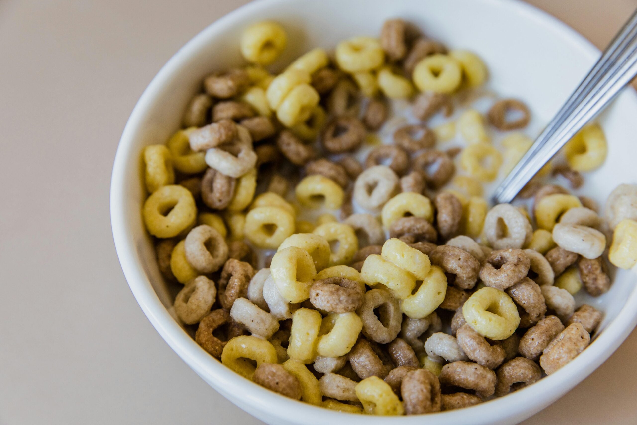 A close-up of a white bowl filled with multicolored, ring-shaped cereal and milk, with a metal spoon resting inside the bowl.