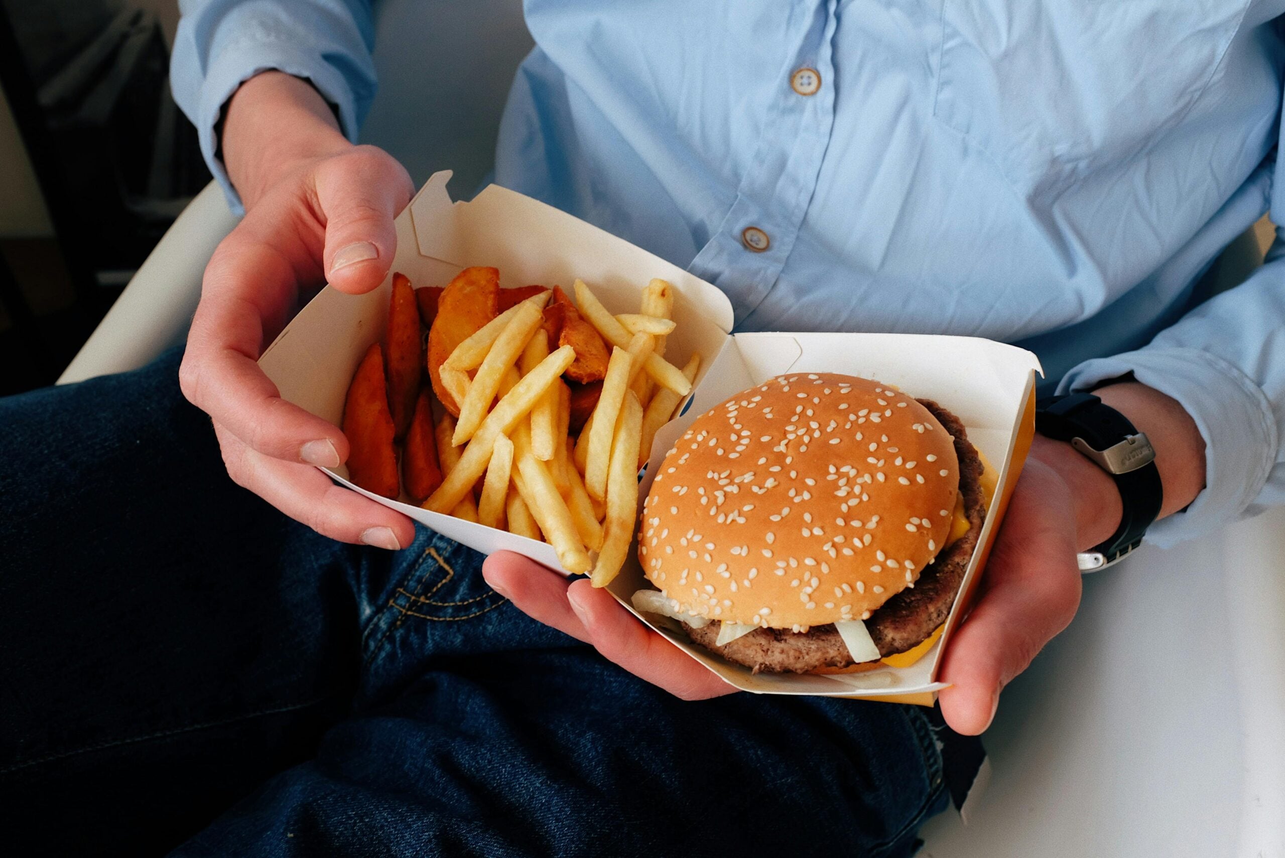 A person in a blue shirt holds a fast food meal on their lap, consisting of a cheeseburger, French fries, and potato wedges in a cardboard tray.