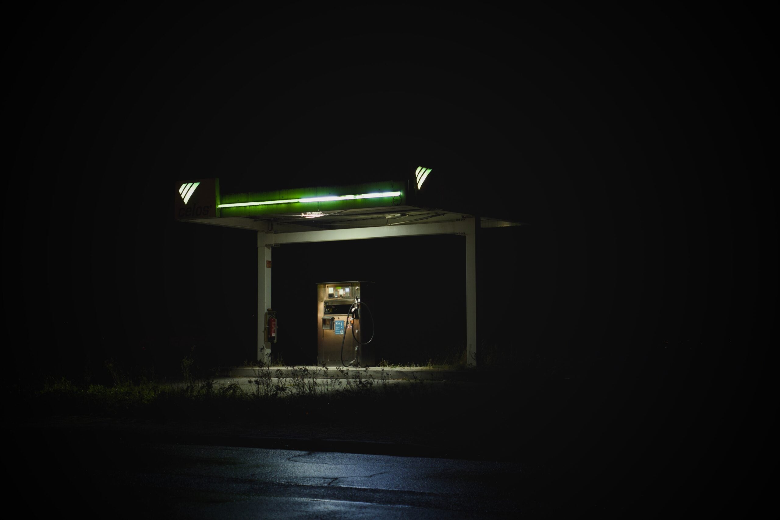 A single, dimly lit gas pump stands under a small canopy at night, surrounded by darkness. The green light above the pump casts a faint glow, while the rest of the area is barely visible.