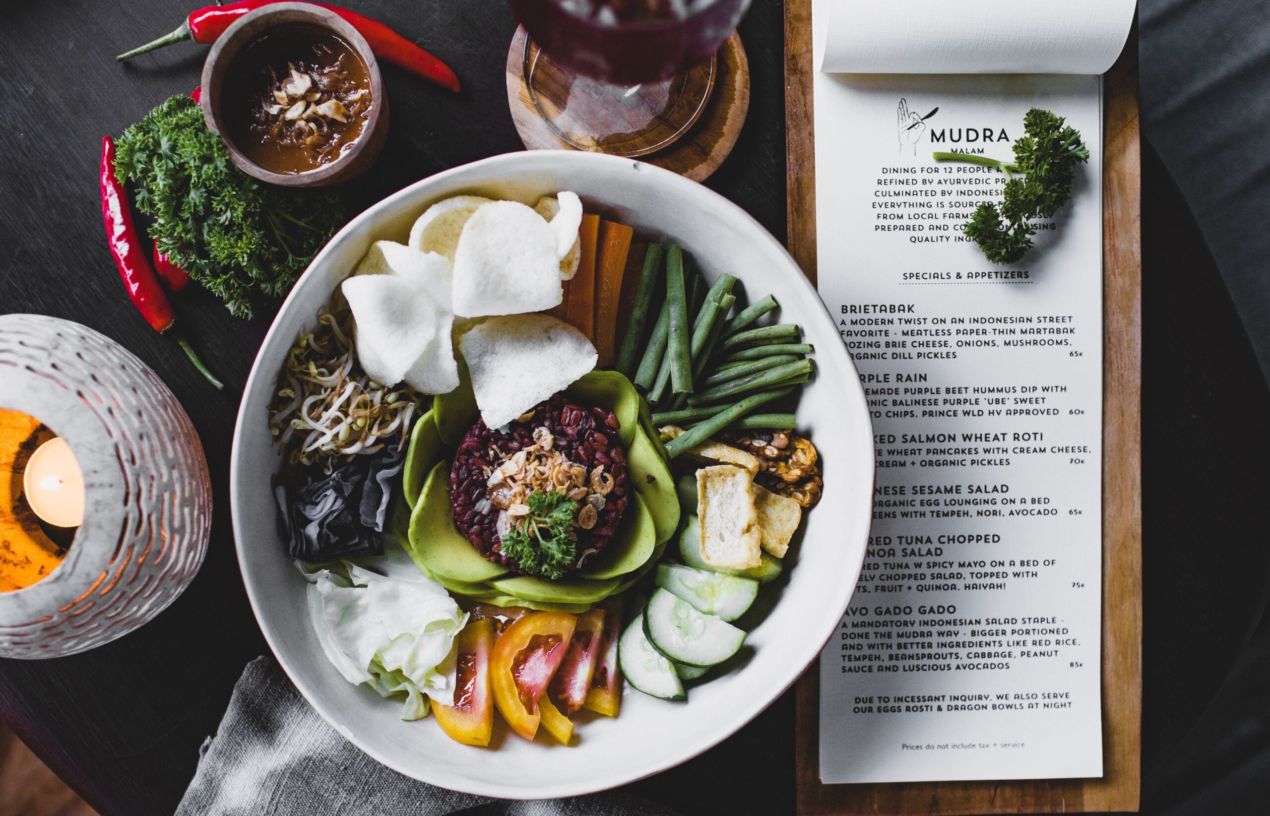 A bowl of colorful vegetables, tofu, rice, and crackers is placed next to a restaurant menu, a small bowl of dipping sauce, and a lit candle on a dark table.