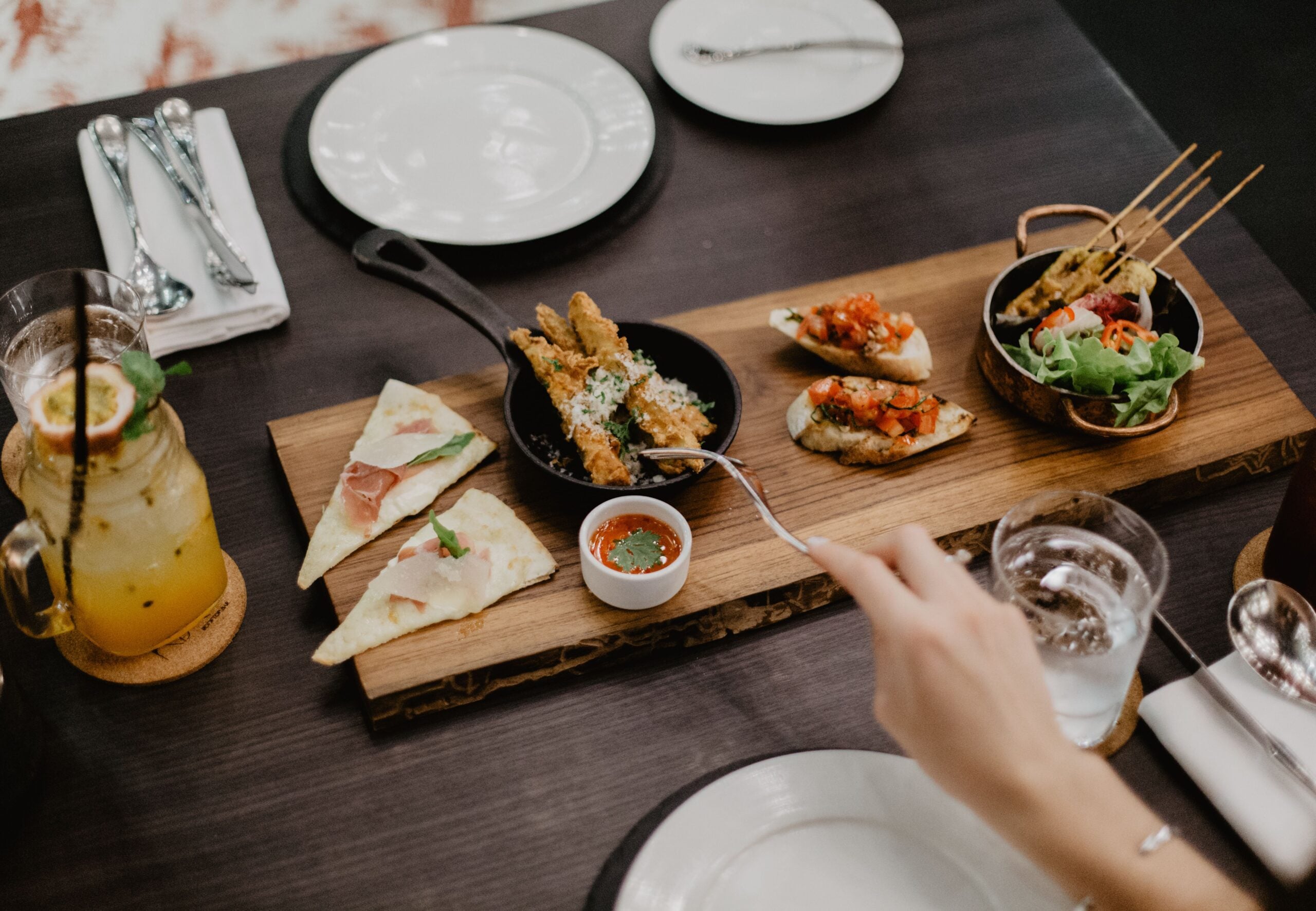 A wooden board holds assorted appetizers including bruschetta, skewers, quesadilla slices, and a small dish of sauce, with a hand reaching in; drinks and plates are set on a dark table.