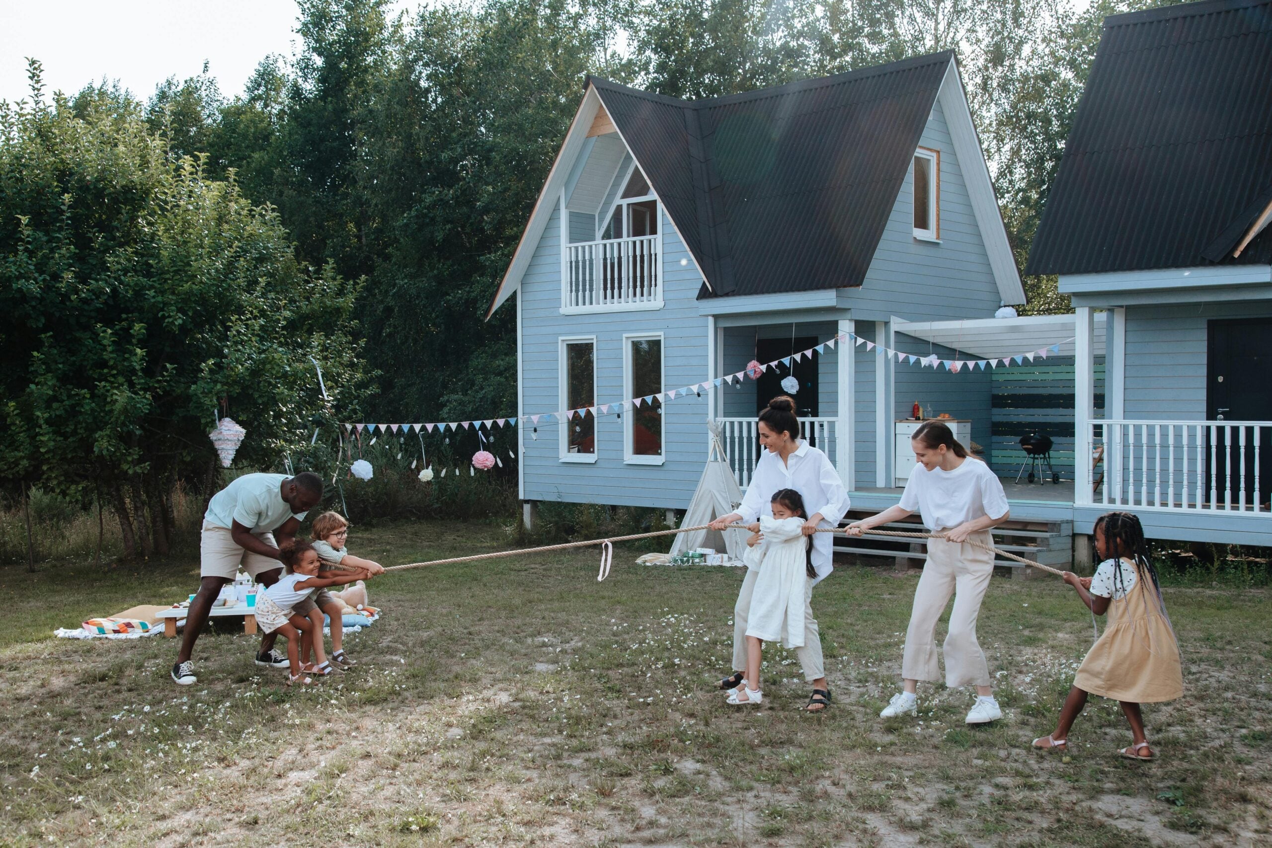 A group of children and adults play tug-of-war on grass in front of two blue houses decorated with bunting. The players are smiling and energetic, enjoying the outdoor activity on a sunny day.