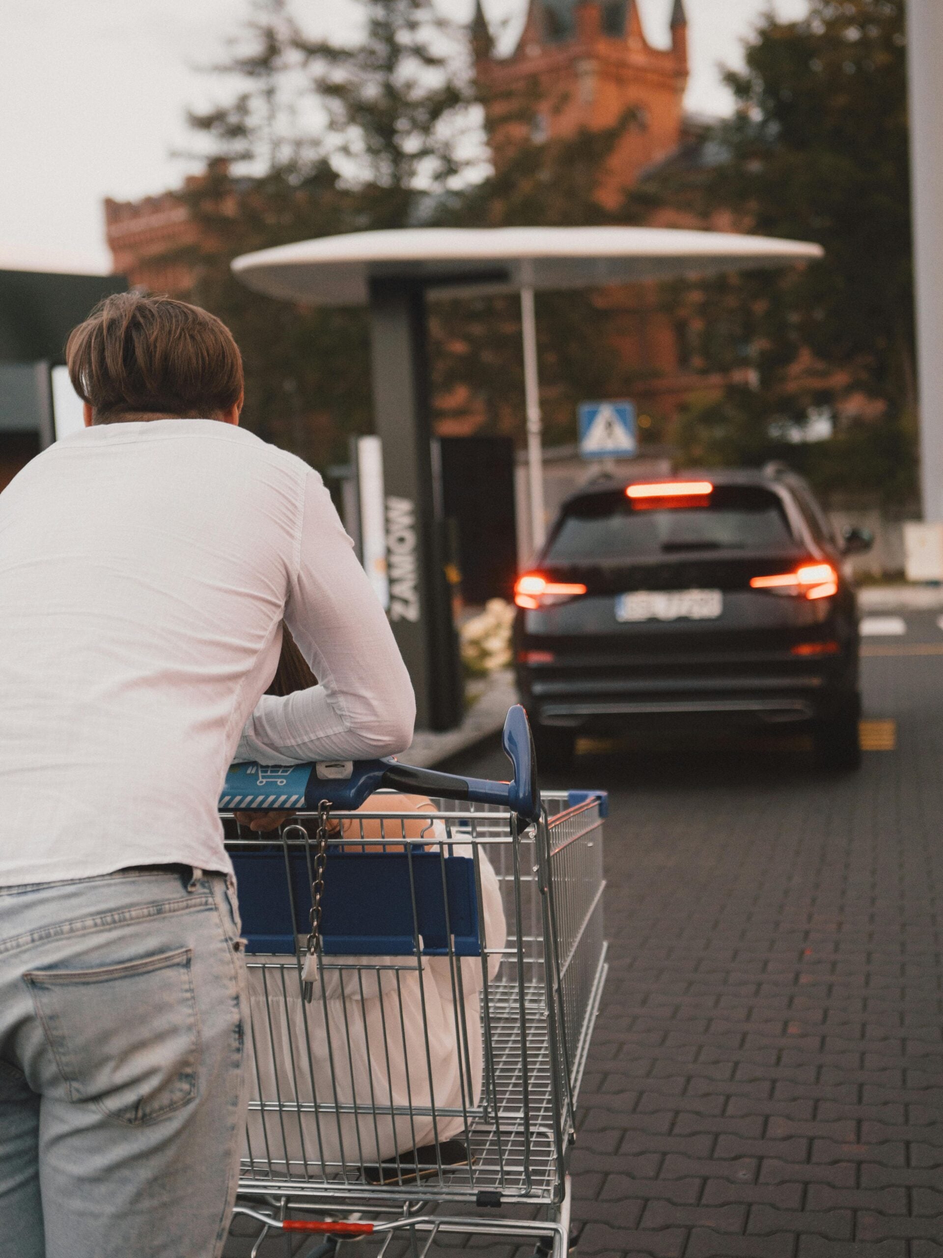 A person in a white shirt pushes a shopping cart outdoors toward a black SUV, which is parked on a paved street near a red brick building and trees.