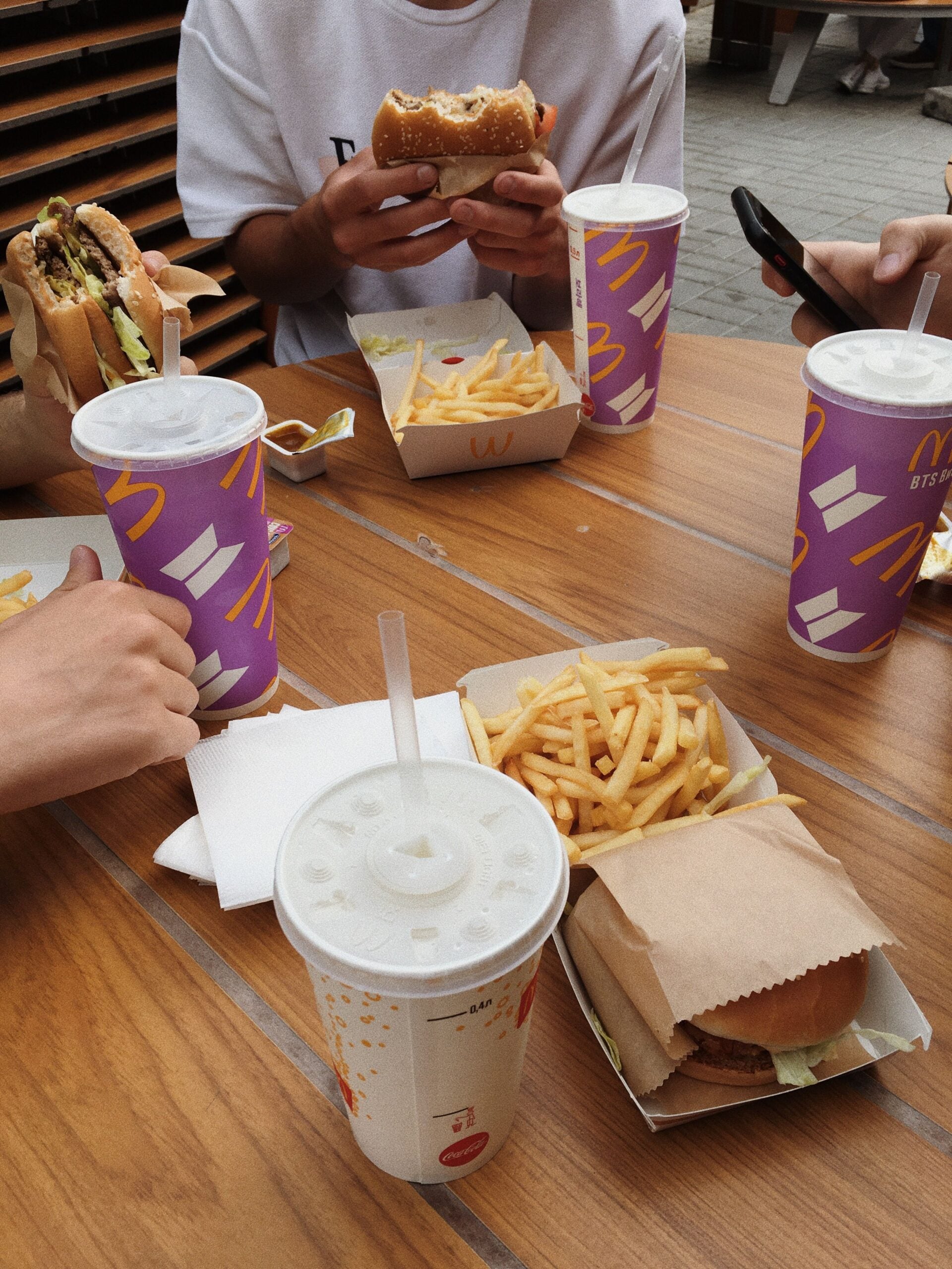 Three people sit at a wooden table with McDonald's food, including burgers, fries, and large drinks in purple cups. One person holds a burger, and another uses a phone. Food wrappers and napkins are scattered on the table.