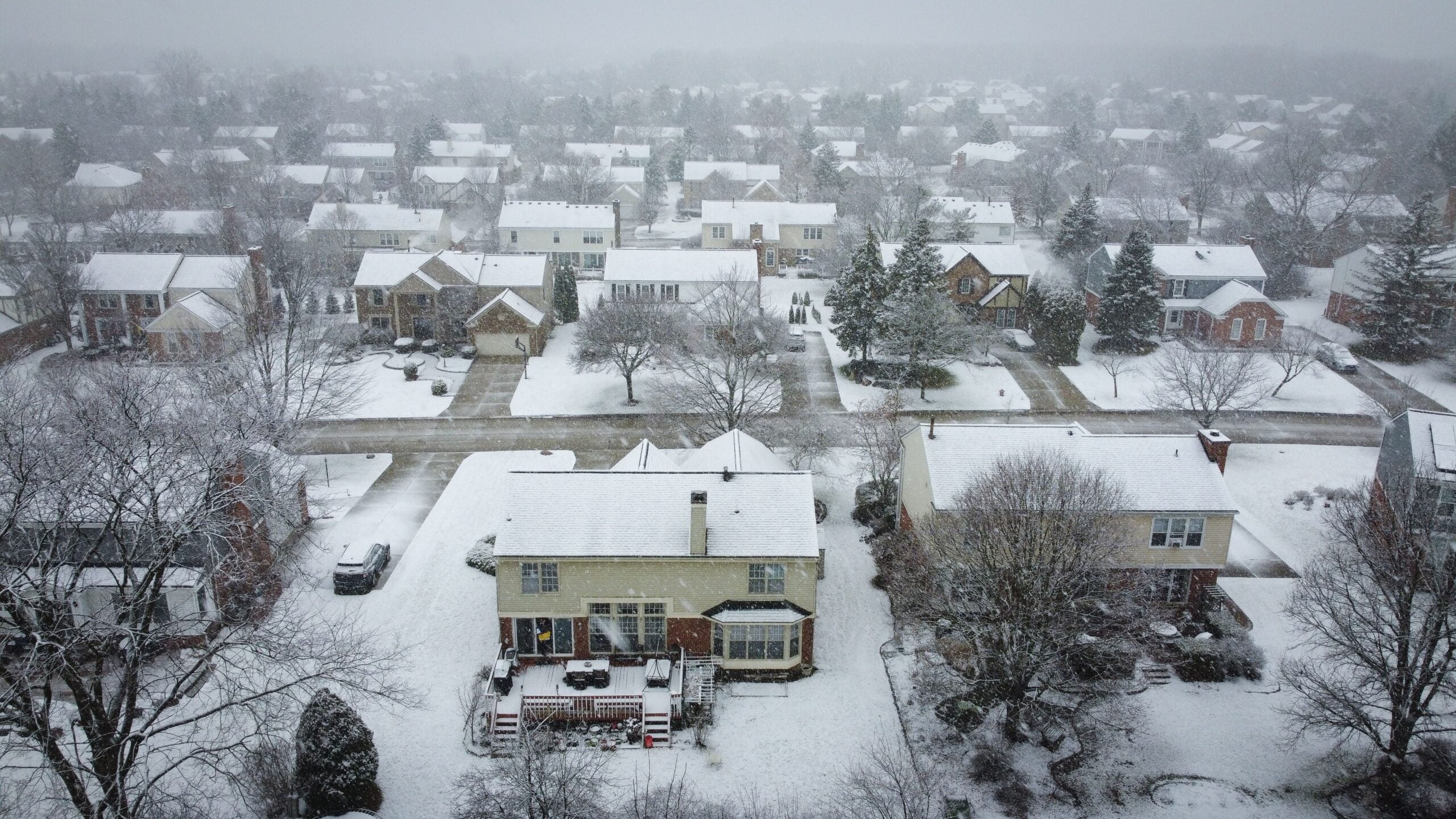 Aerial view of a suburban neighborhood covered in light snow, with houses, leafless trees, and streets blanketed in white on a foggy winter day.