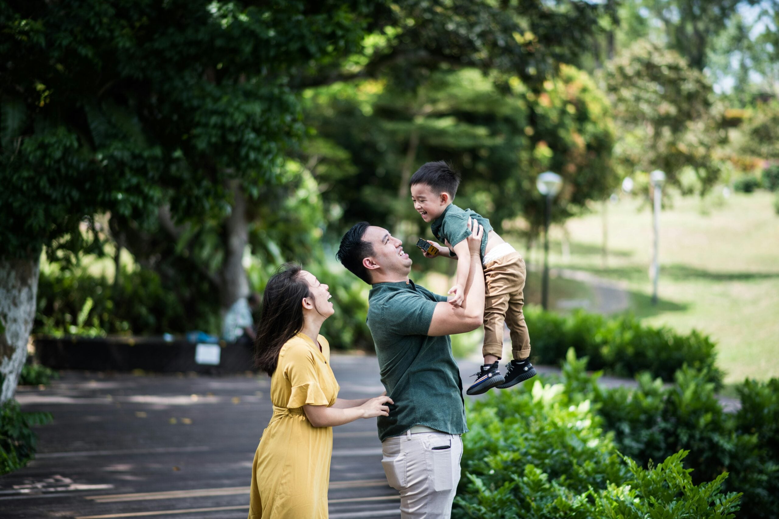 A smiling man lifts a laughing young boy in the air while a woman in a yellow dress looks on, all outdoors in a lush, green park.