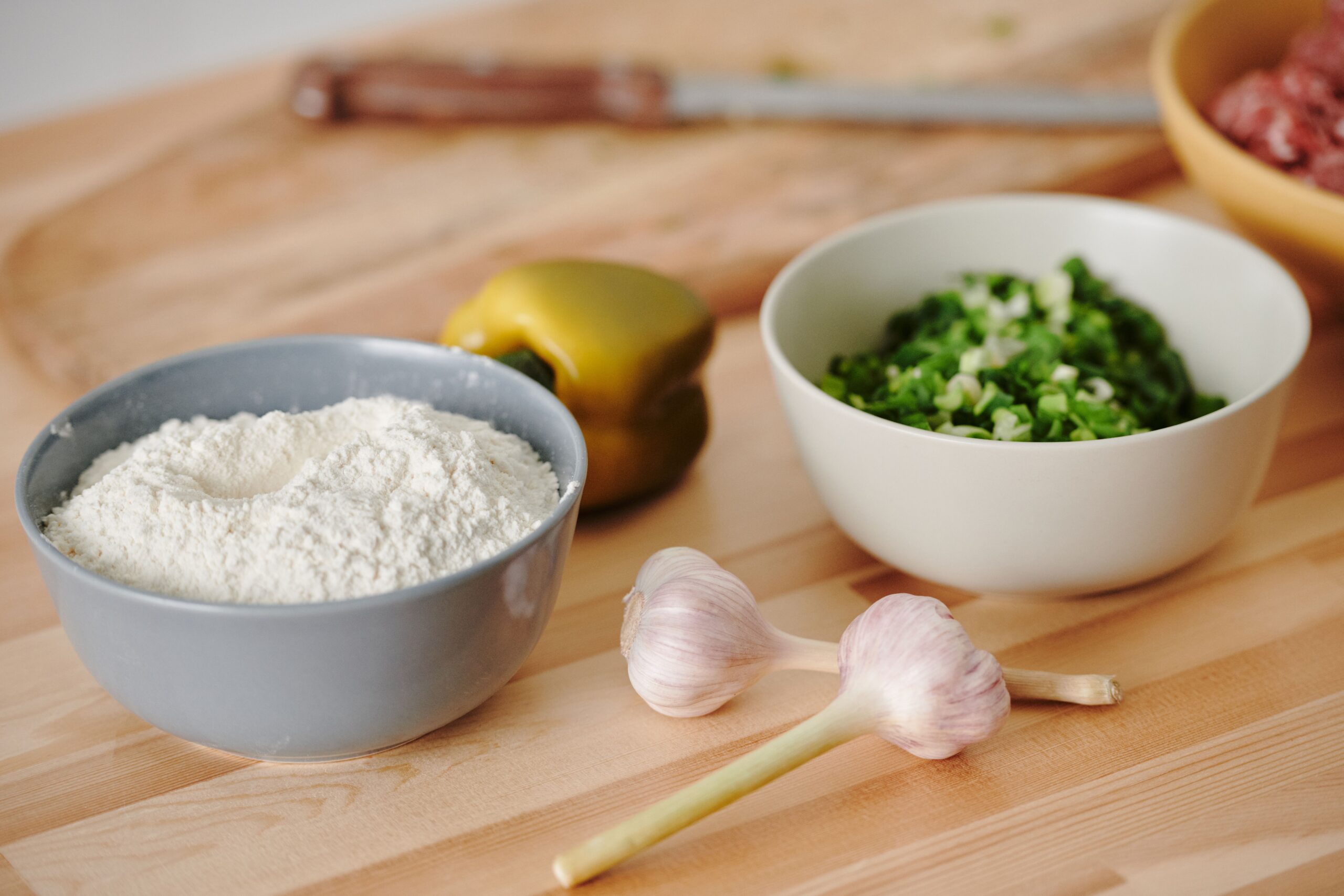 A bowl of flour, a bowl of chopped green vegetables, a yellow bell pepper, two garlic bulbs, and a cutting board with a knife on a wooden countertop.