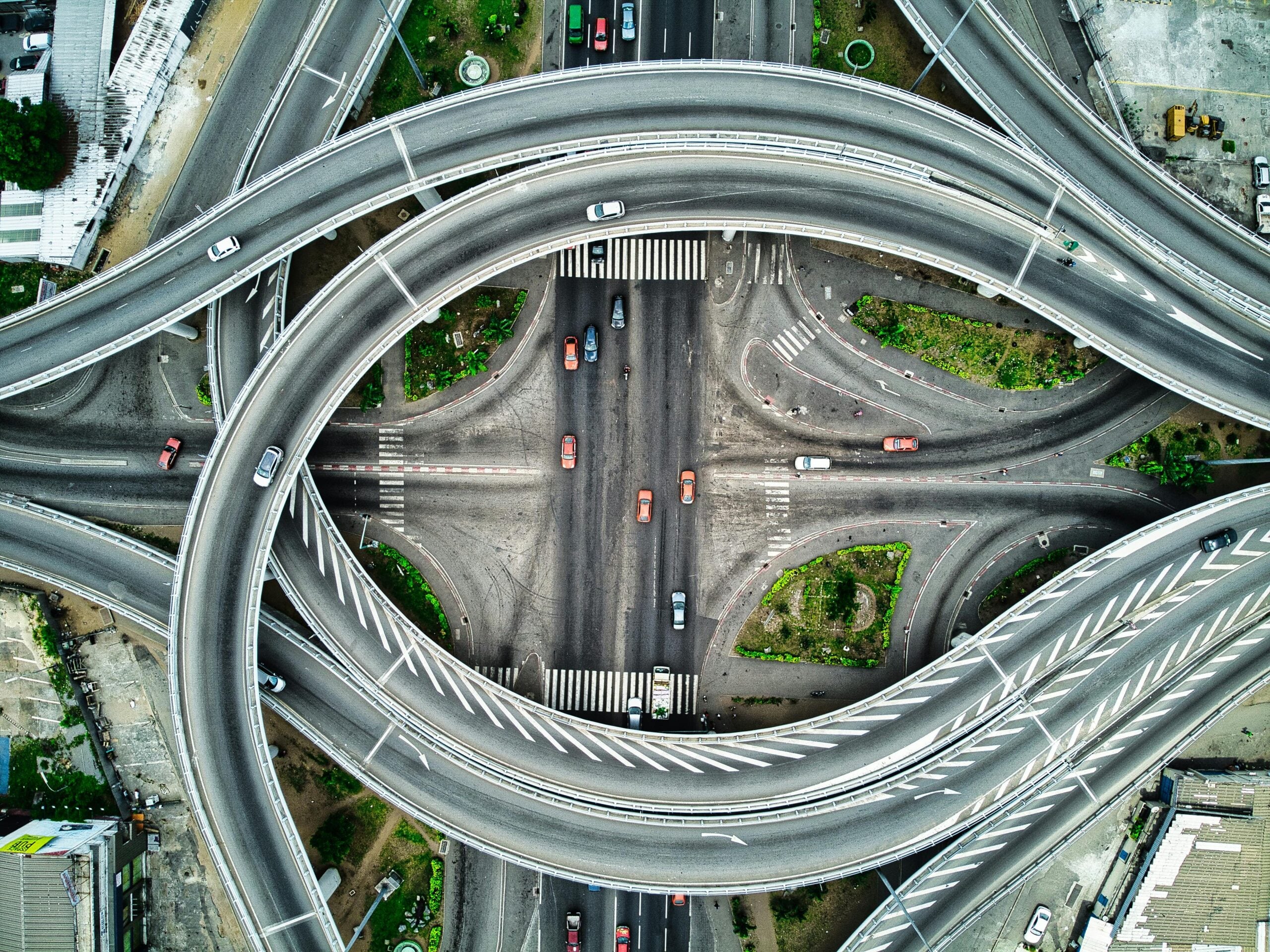 Aerial view of a complex highway interchange with looping overpasses and cars moving on multiple levels above a busy intersection with greenery and crosswalks.