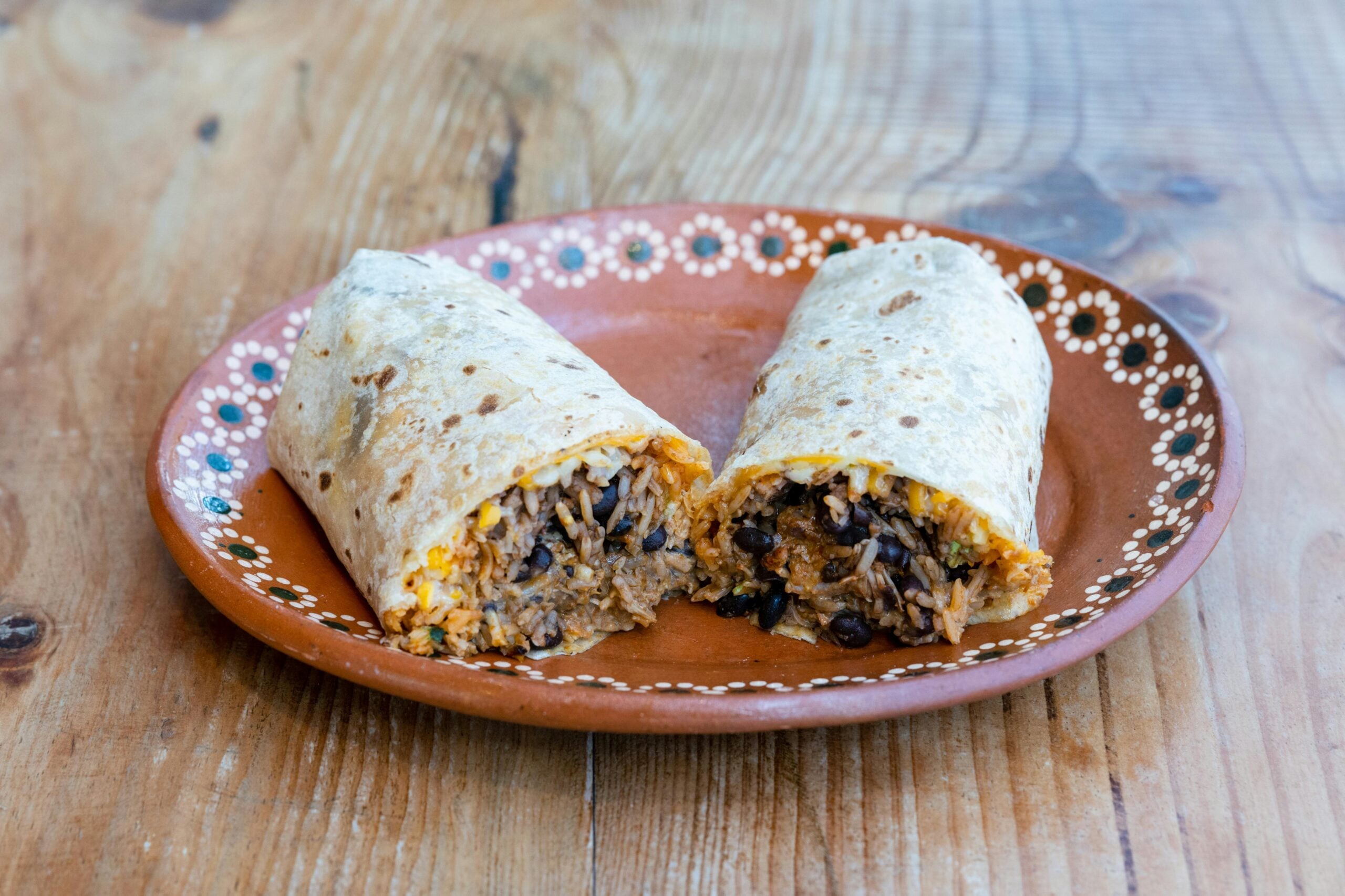 A burrito cut in half, filled with rice, black beans, and cheese, is served on a decorative brown plate placed on a wooden table.