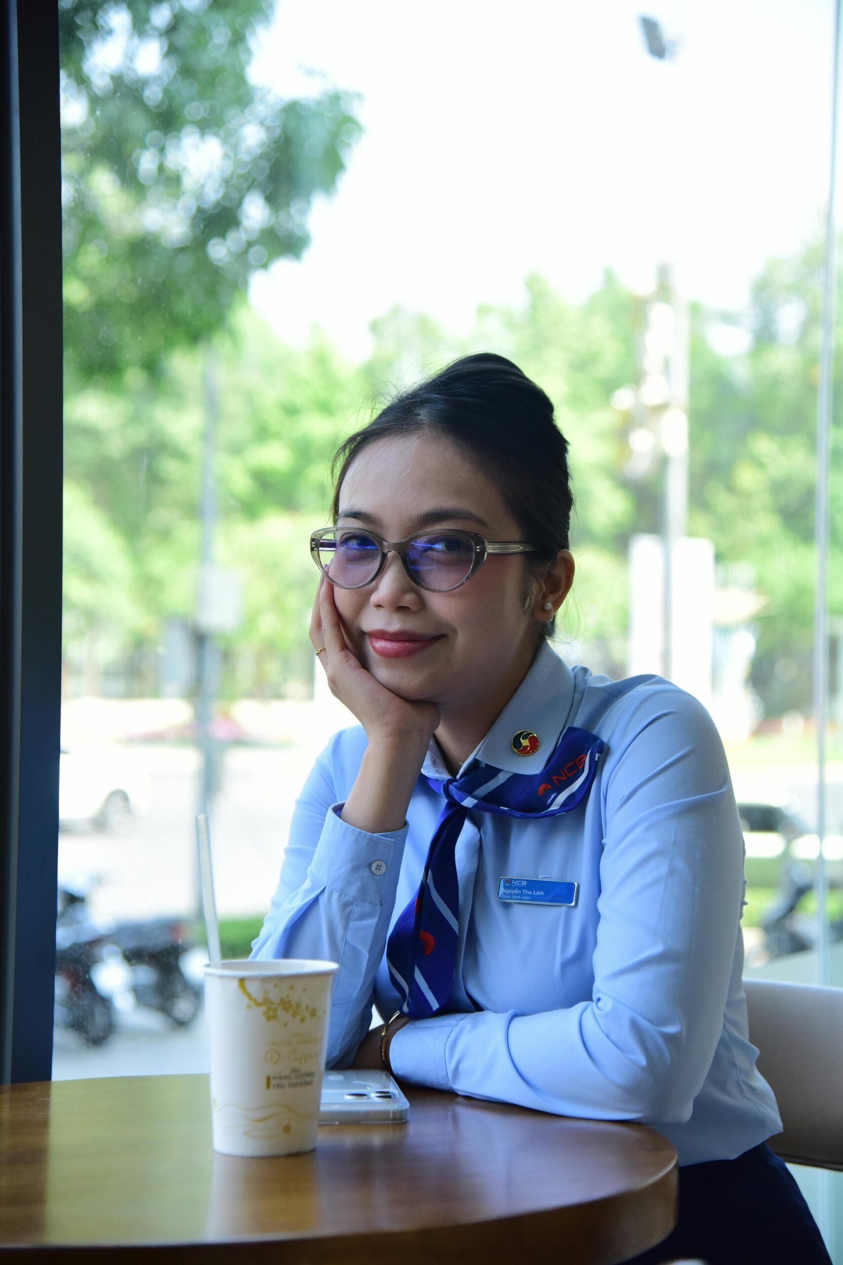 A woman wearing glasses and a light blue uniform with a patterned necktie sits at a table by a window, smiling with her chin resting on her hand. There is a drink cup and a phone on the table.