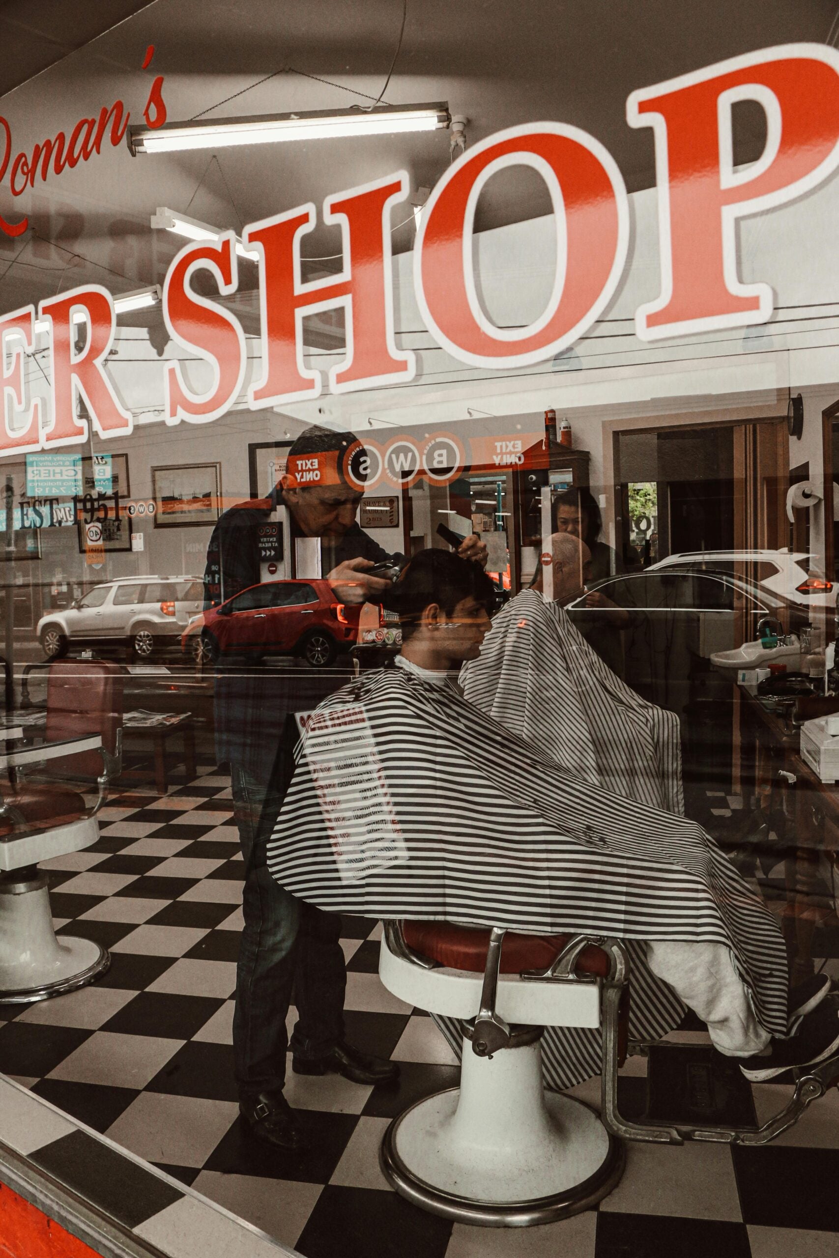 A barber cuts a client's hair inside a barbershop with black-and-white checkered floor, as seen through the shop window with red and white lettering. Cars and buildings are reflected in the glass.