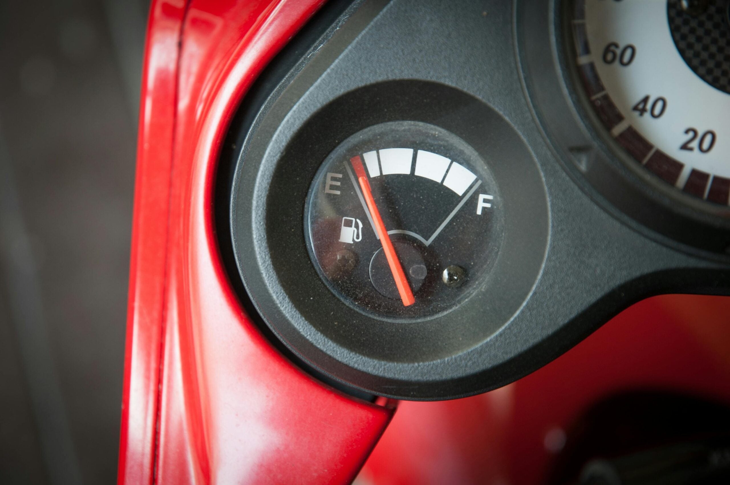 Close-up of a vehicle fuel gauge showing the needle pointing to empty, indicating low fuel. The gauge is set in a black dashboard with part of a red surface visible.