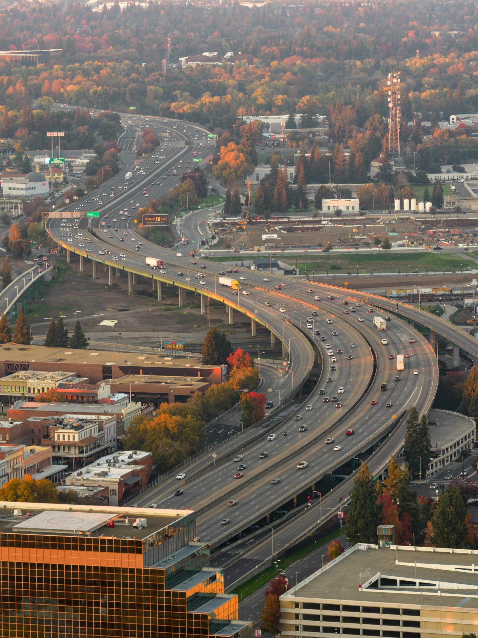 Aerial view of a multi-lane highway with cars and trucks, surrounded by buildings, trees, and construction areas during autumn. Trees display colorful foliage in shades of green, yellow, and orange.
