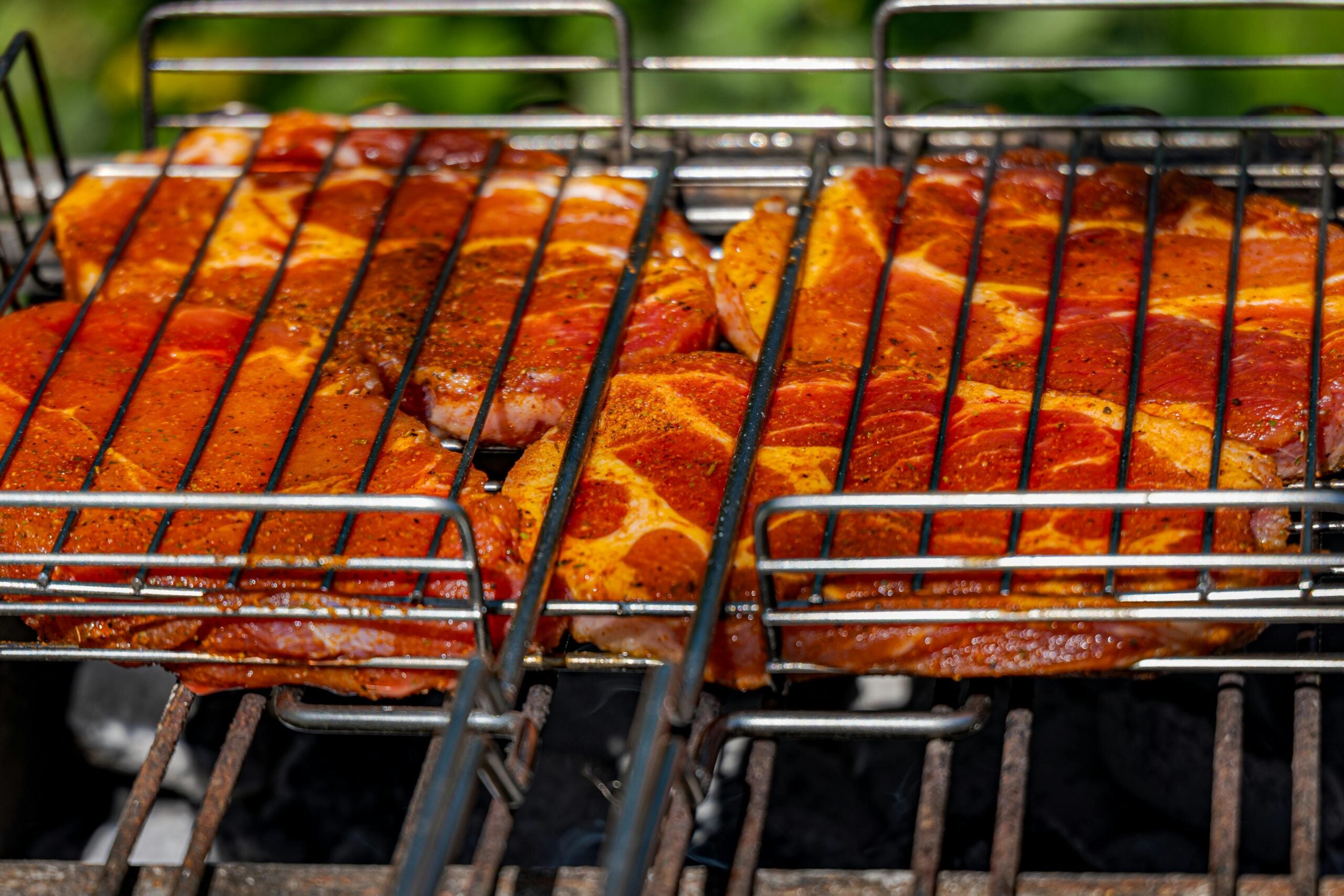 Close-up of seasoned meat slices held in metal grilling racks, cooking over a charcoal grill with blurred greenery in the background.