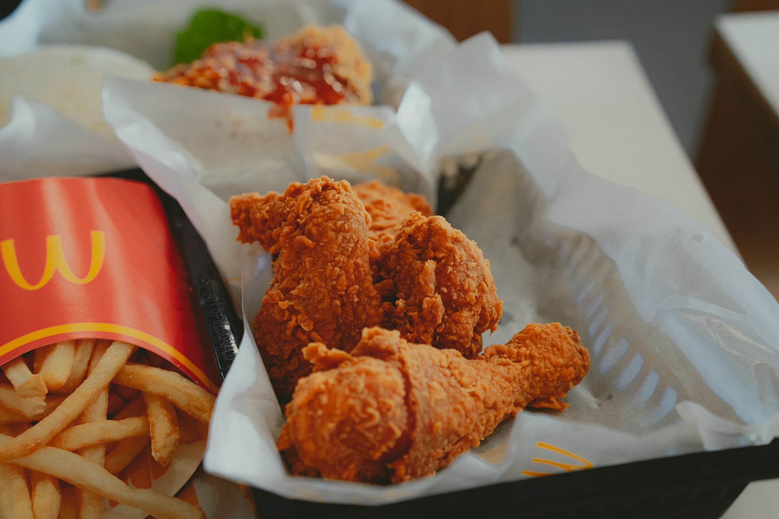 Crispy fried chicken drumsticks in a tray beside a container of McDonald's French fries, with another piece of fried chicken and lettuce in the background.
