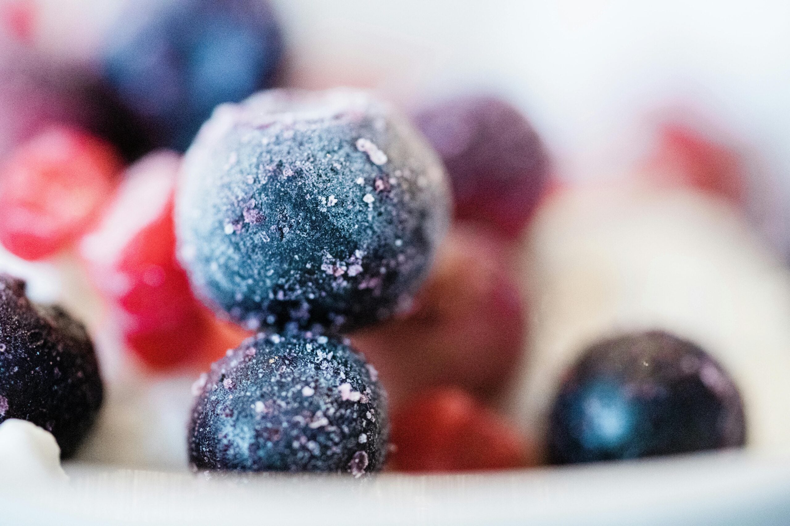 Close-up of assorted frozen berries, including blueberries and red berries, covered in frost, resting on a light surface with a soft, blurred background.