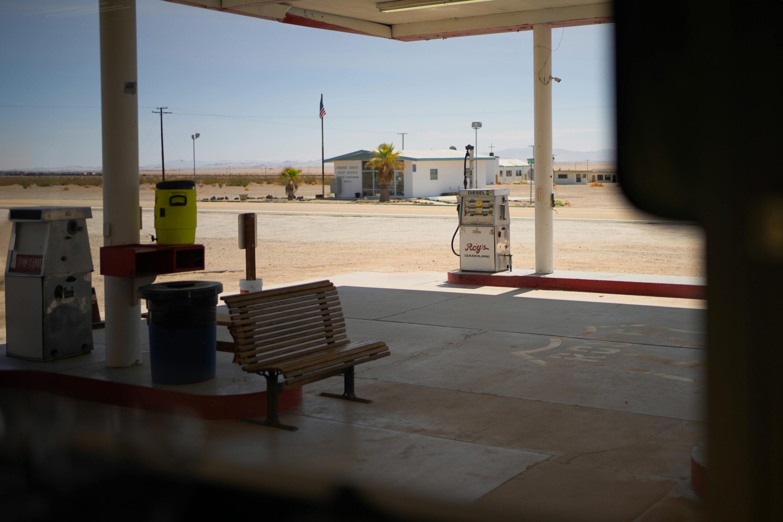 A mostly empty gas station forecourt with a wooden bench, a single vintage gas pump, and a trash can. In the background, there’s a small building, open land, and a distant American flag.