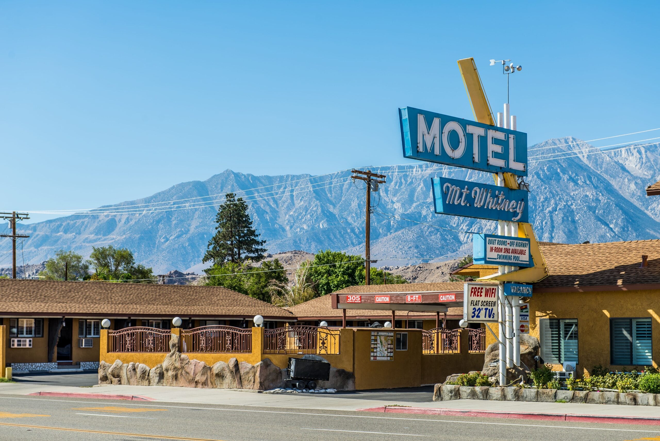 A retro roadside motel with a large blue "MOTEL" sign reading "Mt. Whitney" stands against a backdrop of mountains and clear sky, with brown buildings and a parking area in front.