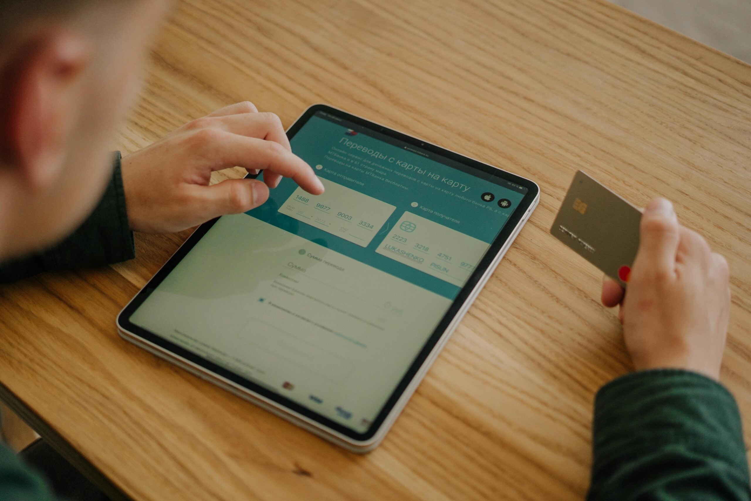 A person uses a tablet for online banking or payments, entering information on the screen while holding a credit card in their other hand. The scene takes place at a wooden table.