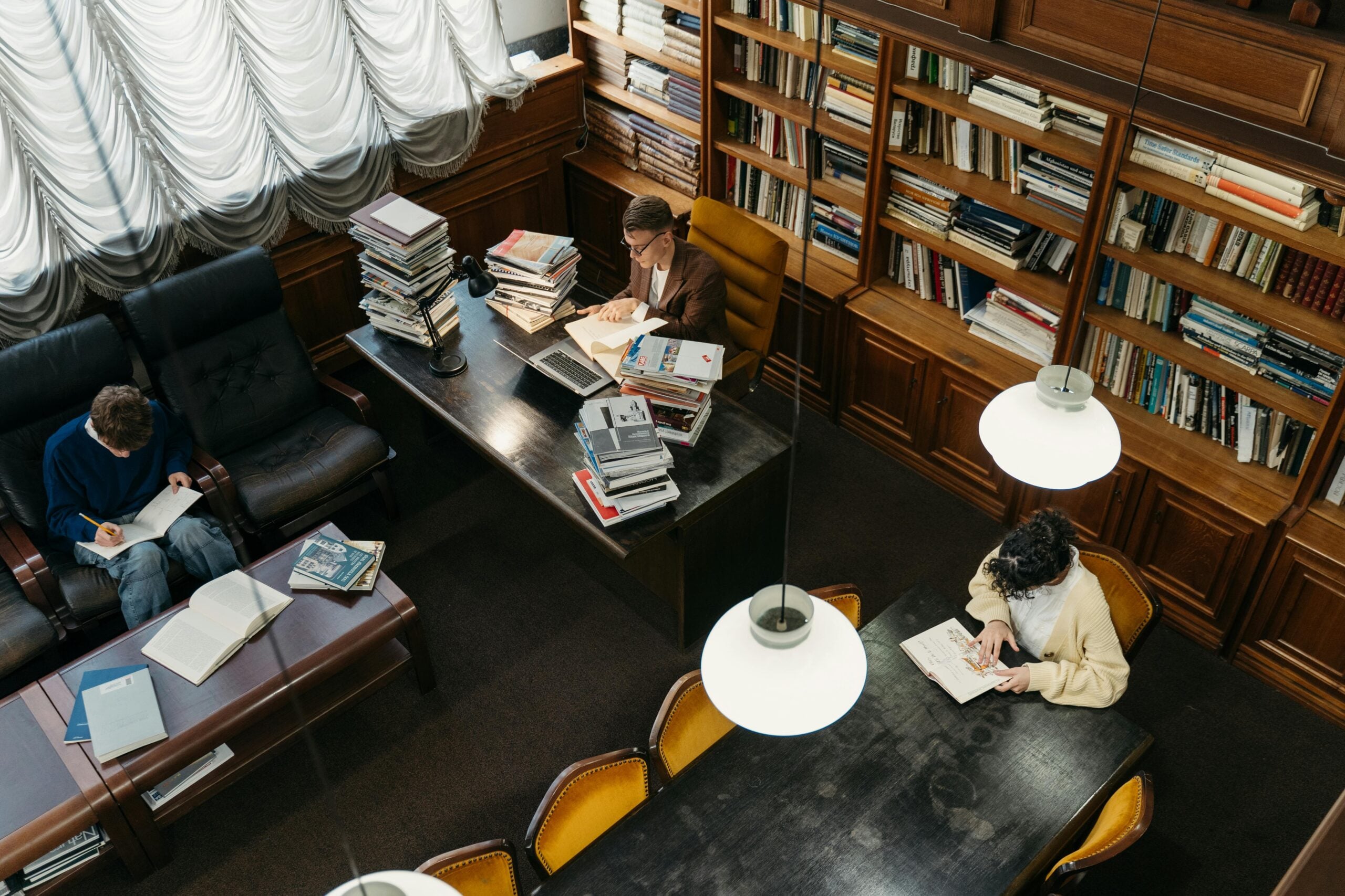 Three people are reading and working at tables in a cozy library filled with bookshelves and stacks of books. Warm lighting hangs above, and the room has armchairs and large windows with draped curtains.