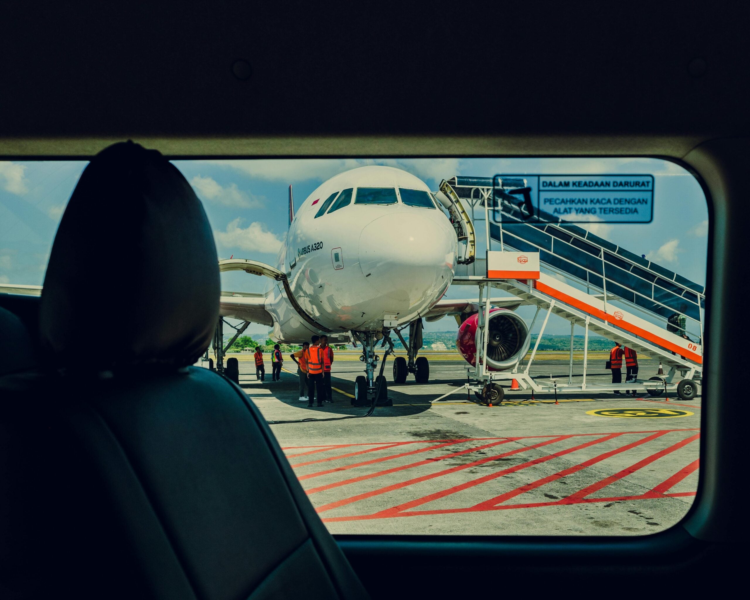 A view from inside a vehicle shows an airplane on the tarmac, with ground crew in orange vests standing near the nose and a boarding staircase positioned at the plane’s door.