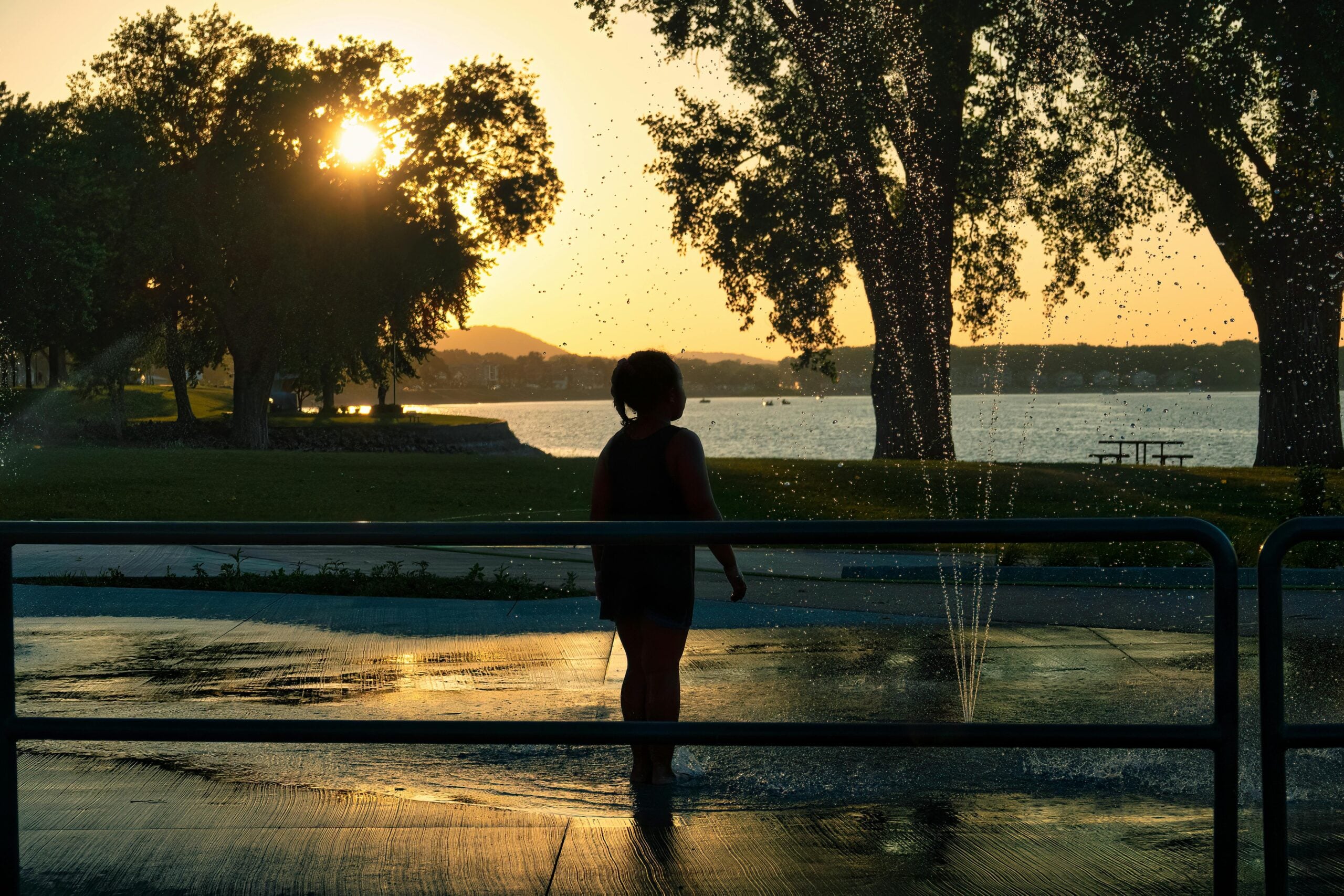 A child stands in the spray of a fountain at sunset, silhouetted against the golden light, with trees, grass, and a lake in the background.